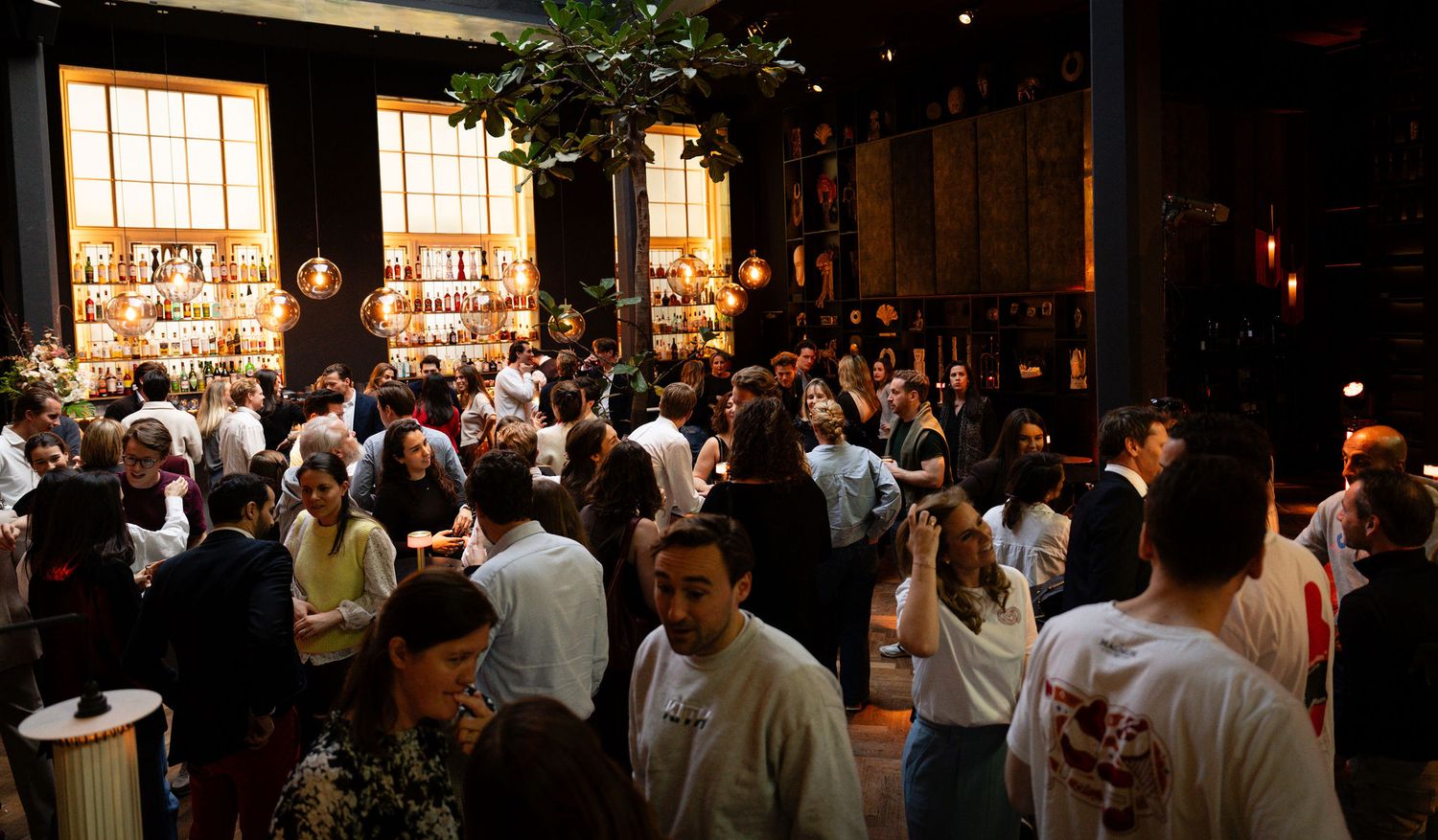 Crowded indoor event with guests mingling near a bar and bookshelves in a warmly lit venue.