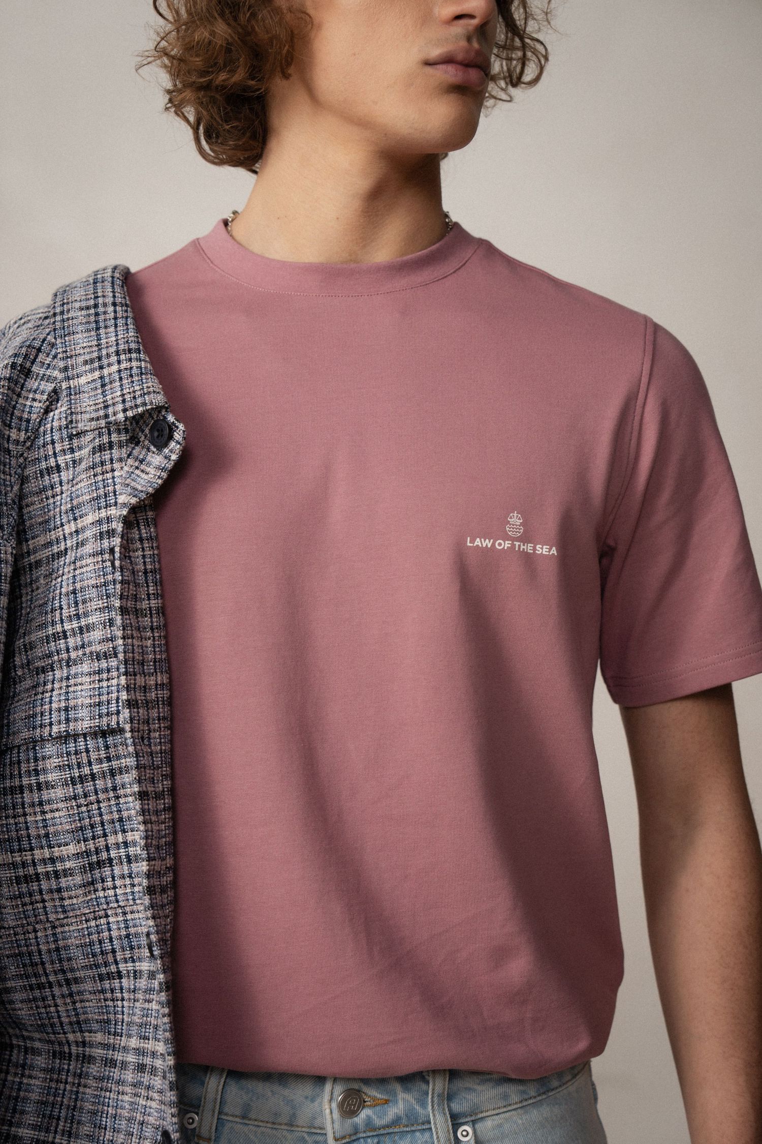 Model wearing a dusty rose colored t-shirt with small text detail and plaid overshirt in studio lighting.