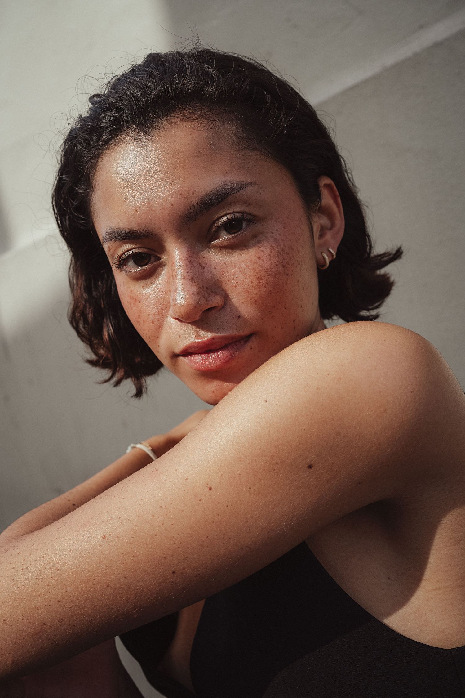 Young woman with freckles and short dark hair poses thoughtfully, wearing a dark top, in natural sunlight.