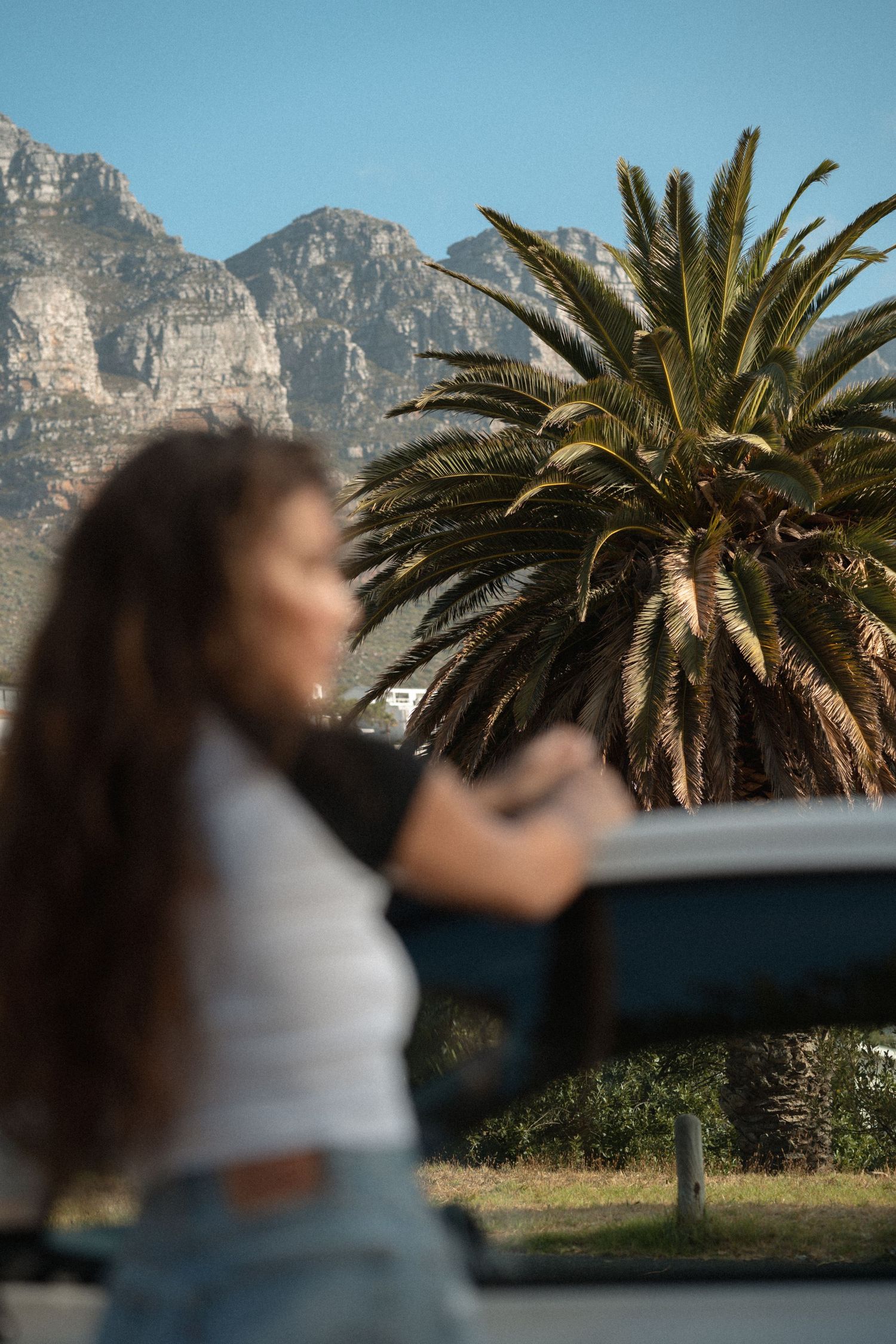 Woman standing by a car with a palm tree and dramatic mountain backdrop on a sunny day.