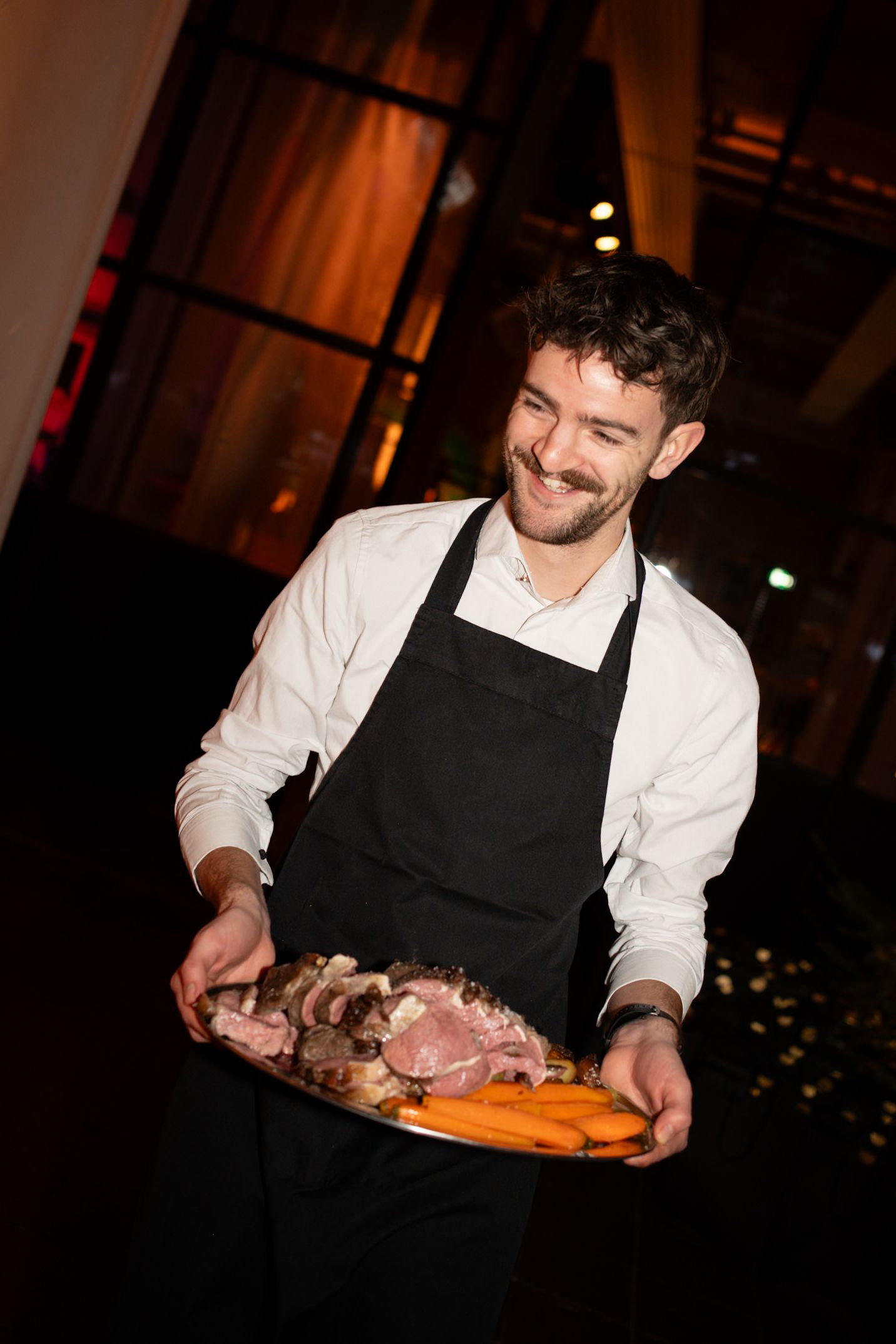Smiling waiter in black apron carrying a large platter of carved roasted meat and carrots at a catered event.