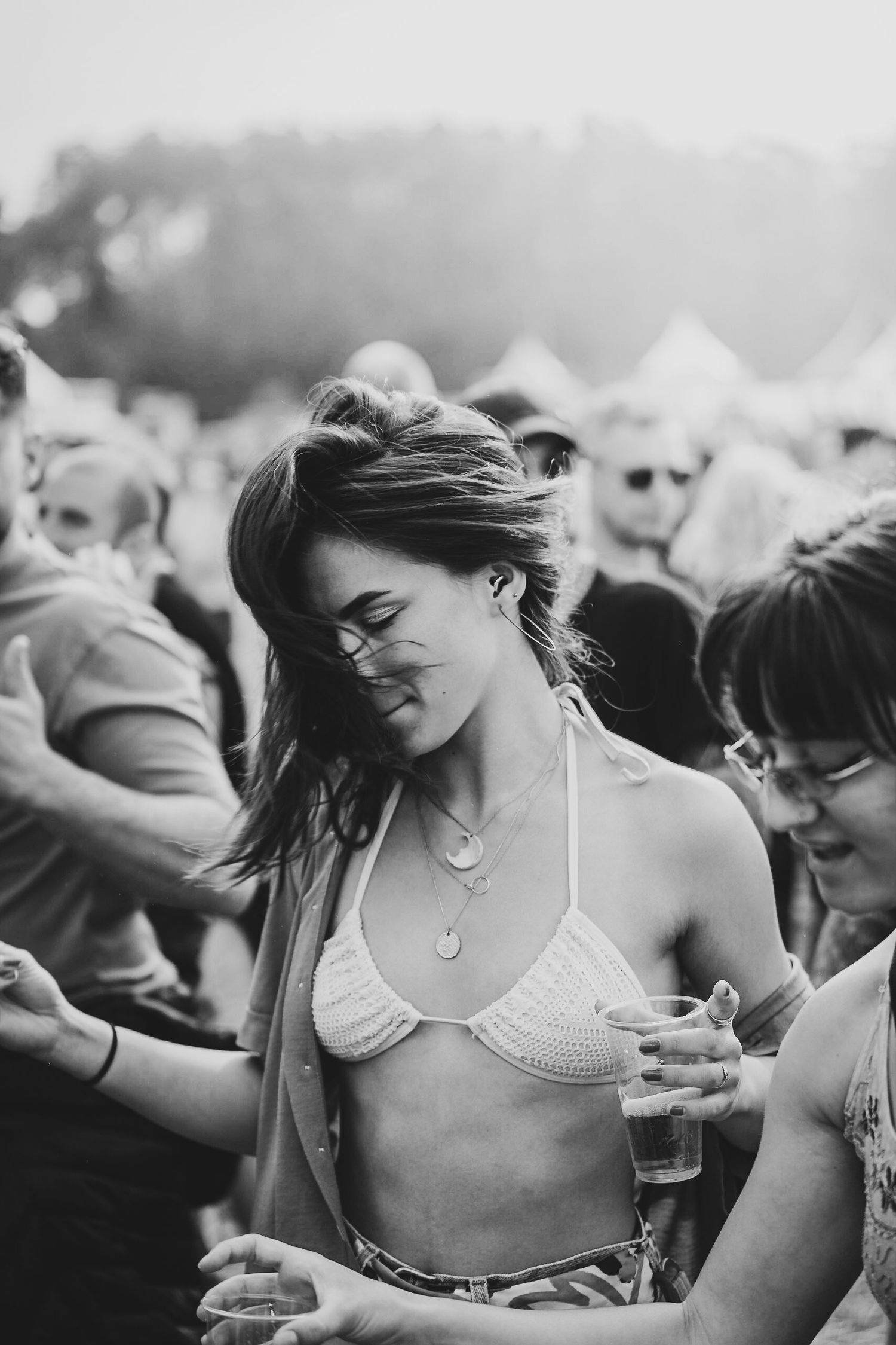 Black and white photo of a woman dancing joyfully at an outdoor music festival, holding a drink in a crowd.