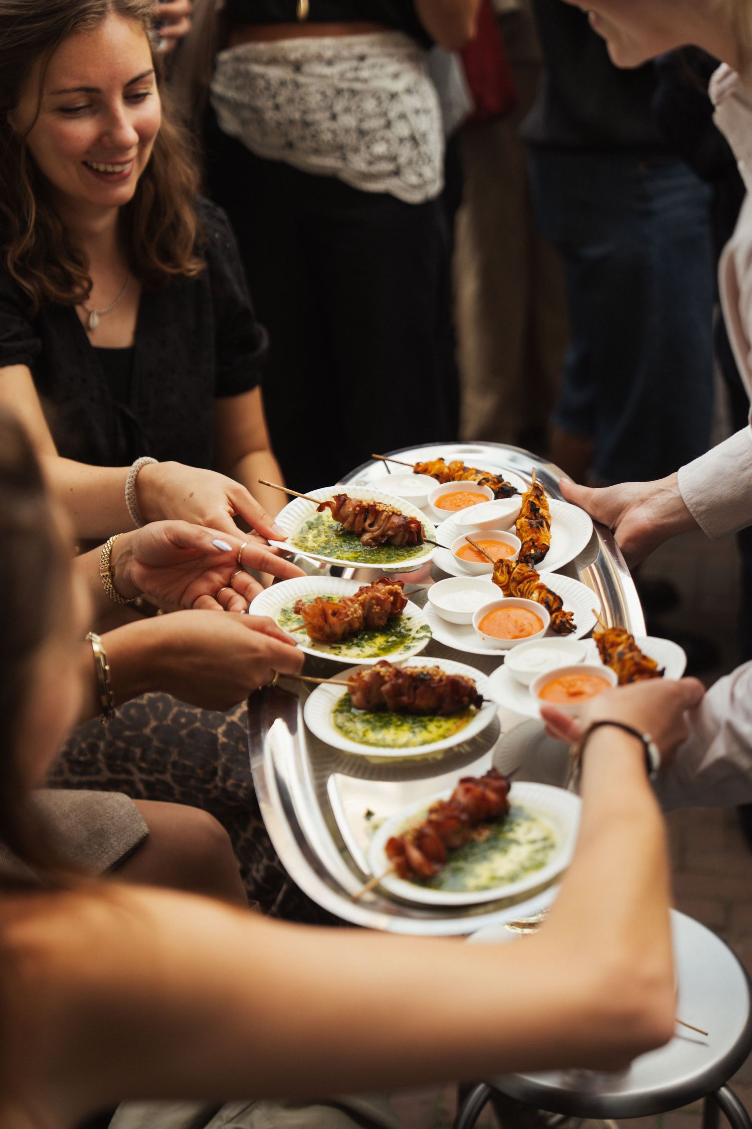 People sharing skewered appetizers on silver tray at social gathering, with dipping sauces and smiling guests.