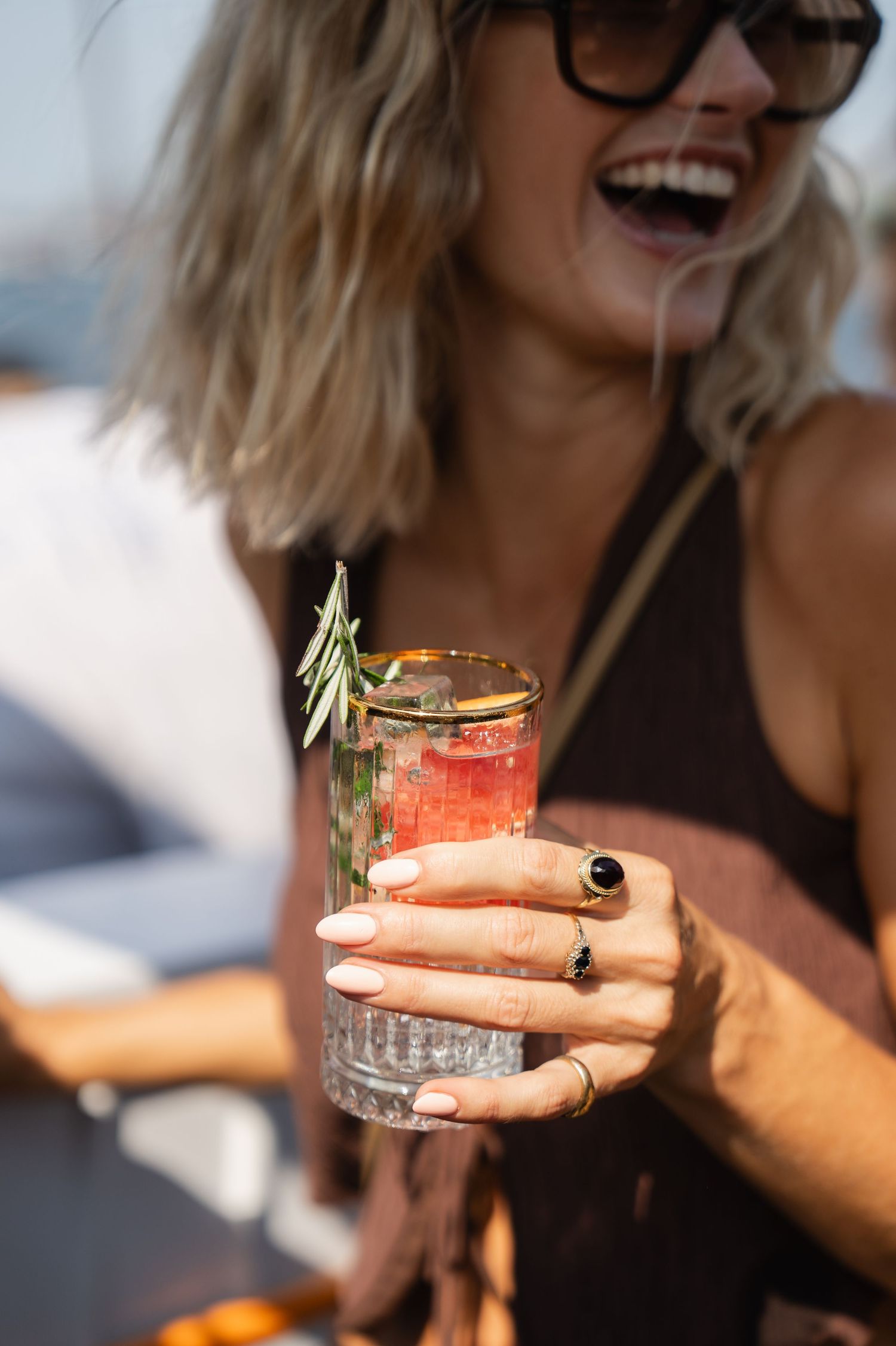 A laughing blonde woman in sunglasses holds a garnished pink cocktail with rosemary and citrus at an outdoor event.