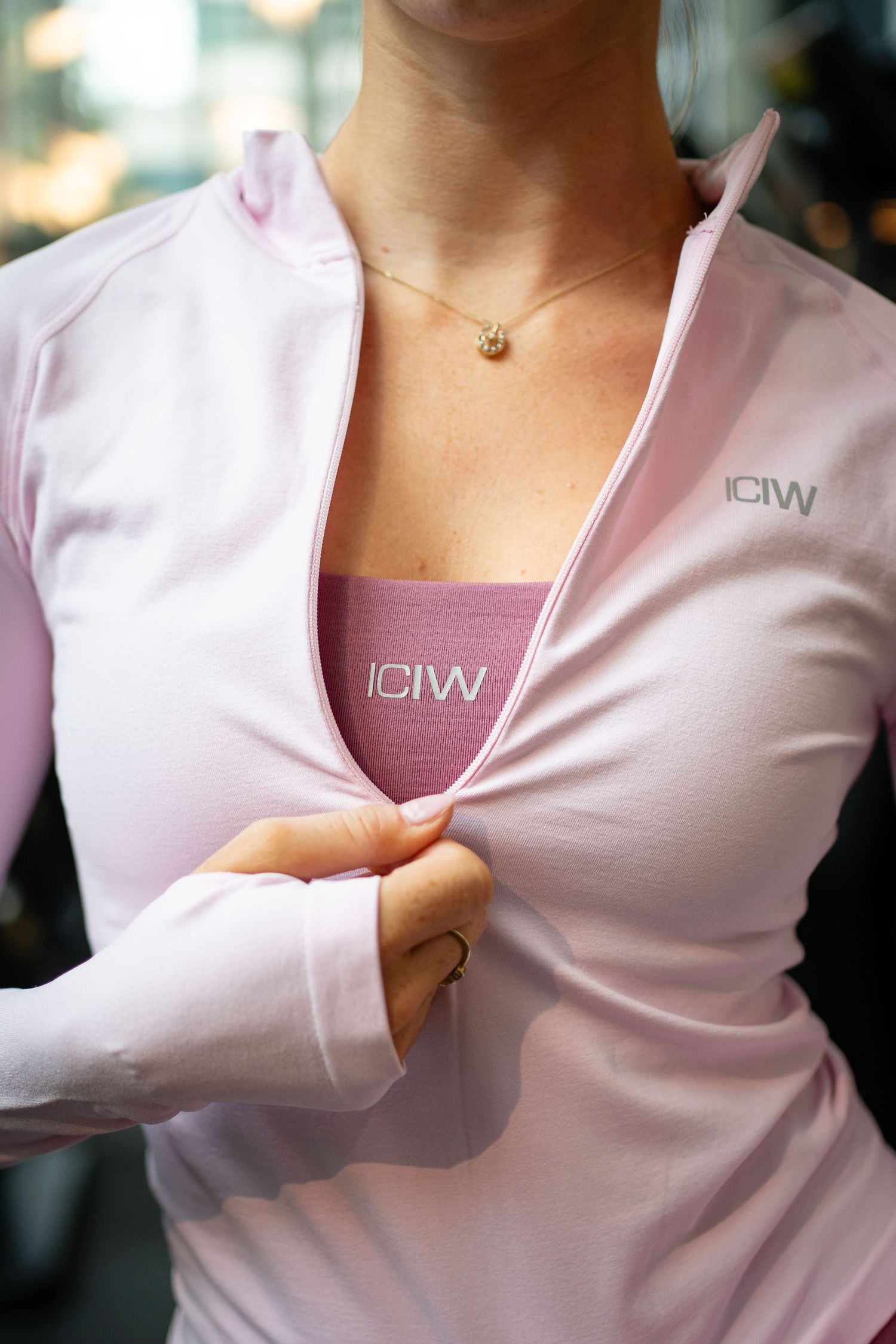 Woman wearing pink ICIW long sleeve zip-up jacket over a mauve ICIW sports bra, with a gold necklace.