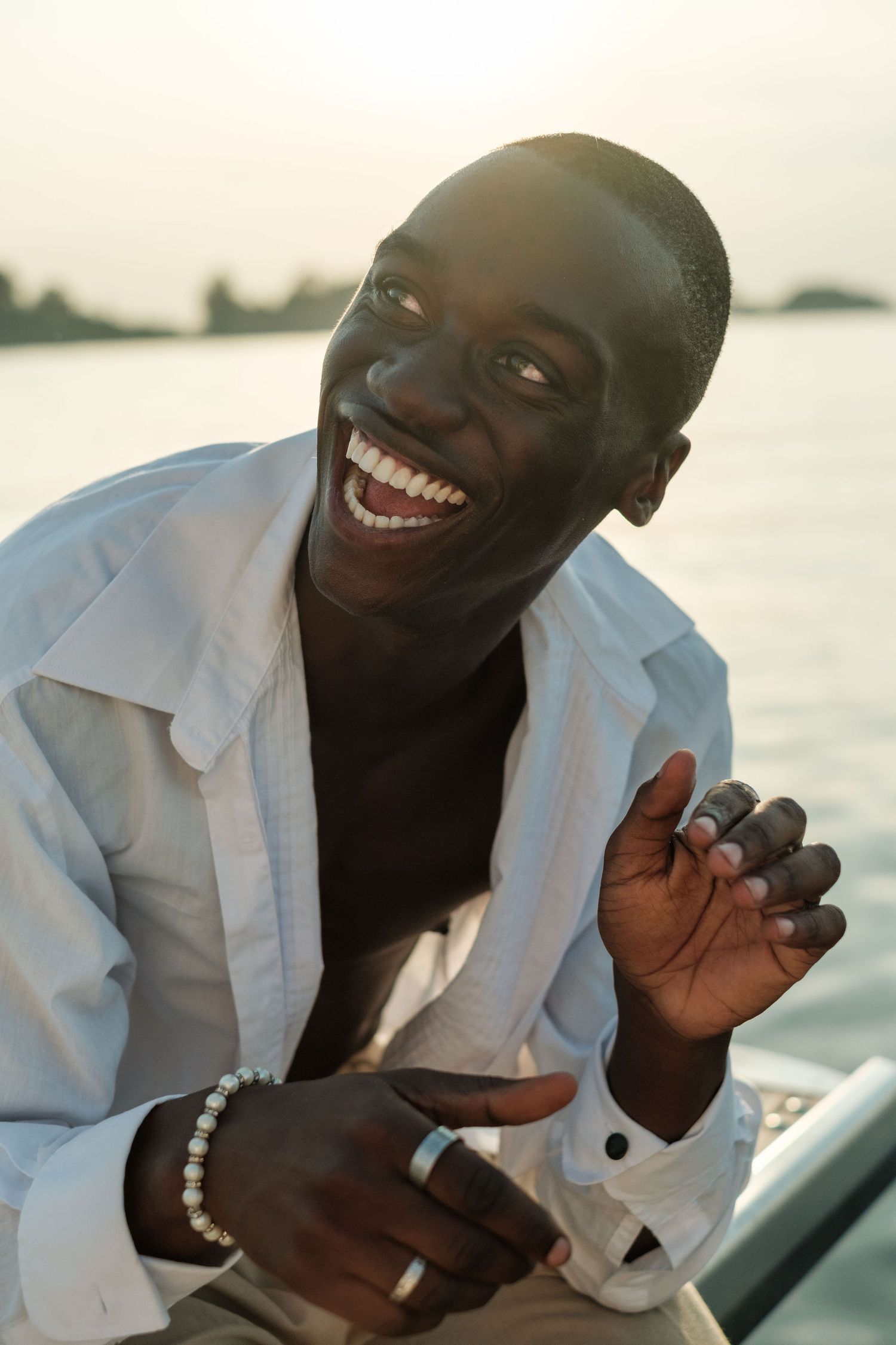 A laughing Black man in a white shirt and pearl bracelet sits on a boat at sunset, showcasing rings on his fingers.