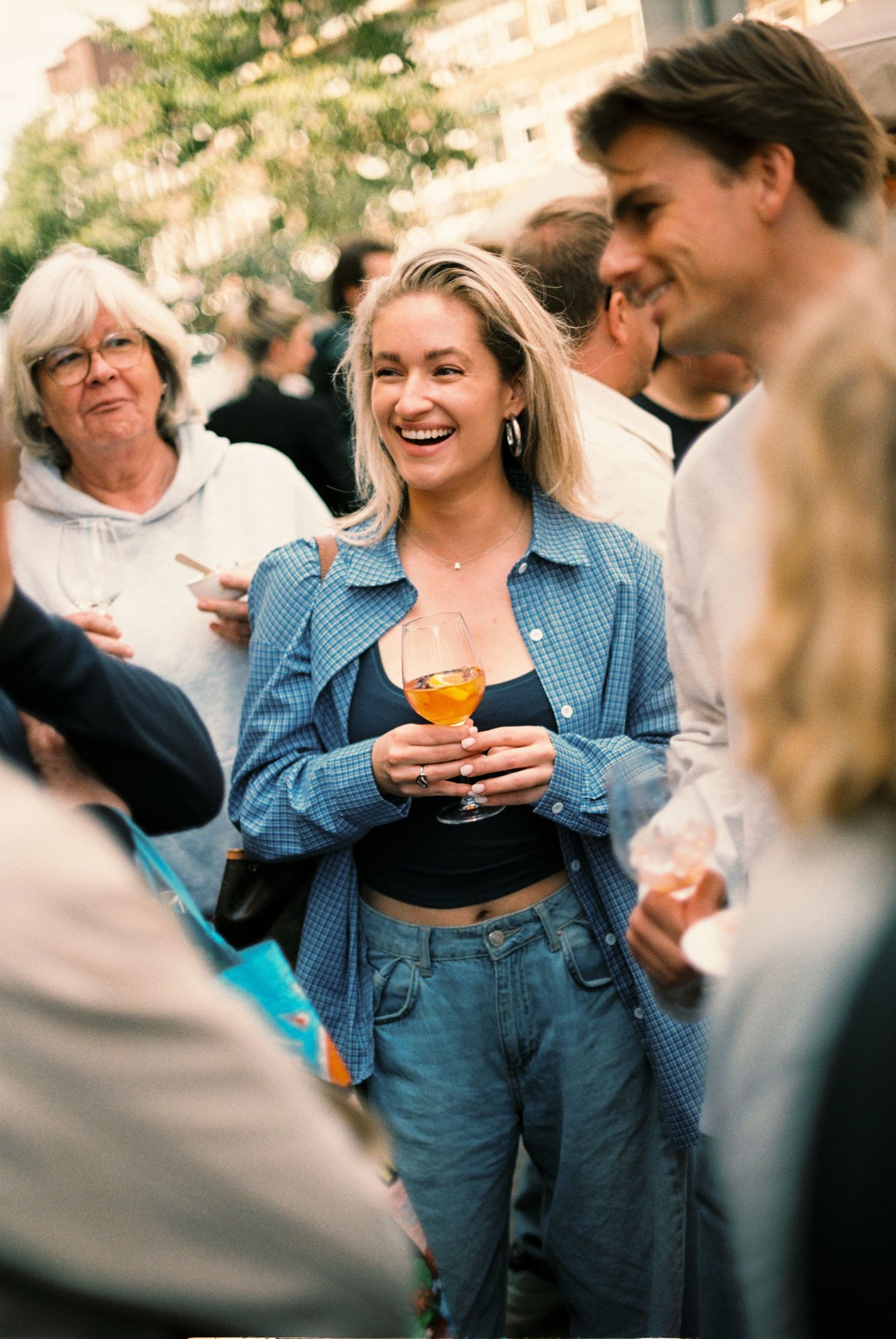Smiling blonde woman in blue denim outfit holding a cocktail at an outdoor social gathering.