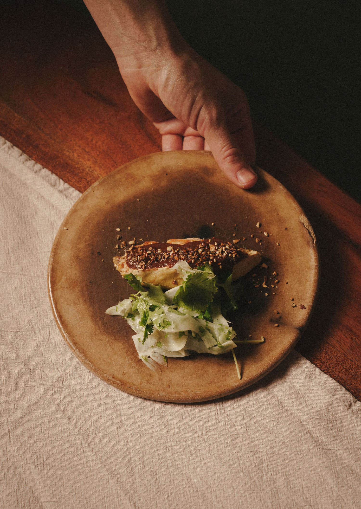 Hand placing a rustic ceramic plate with sesame-crusted fish and fresh green salad on a linen tablecloth.