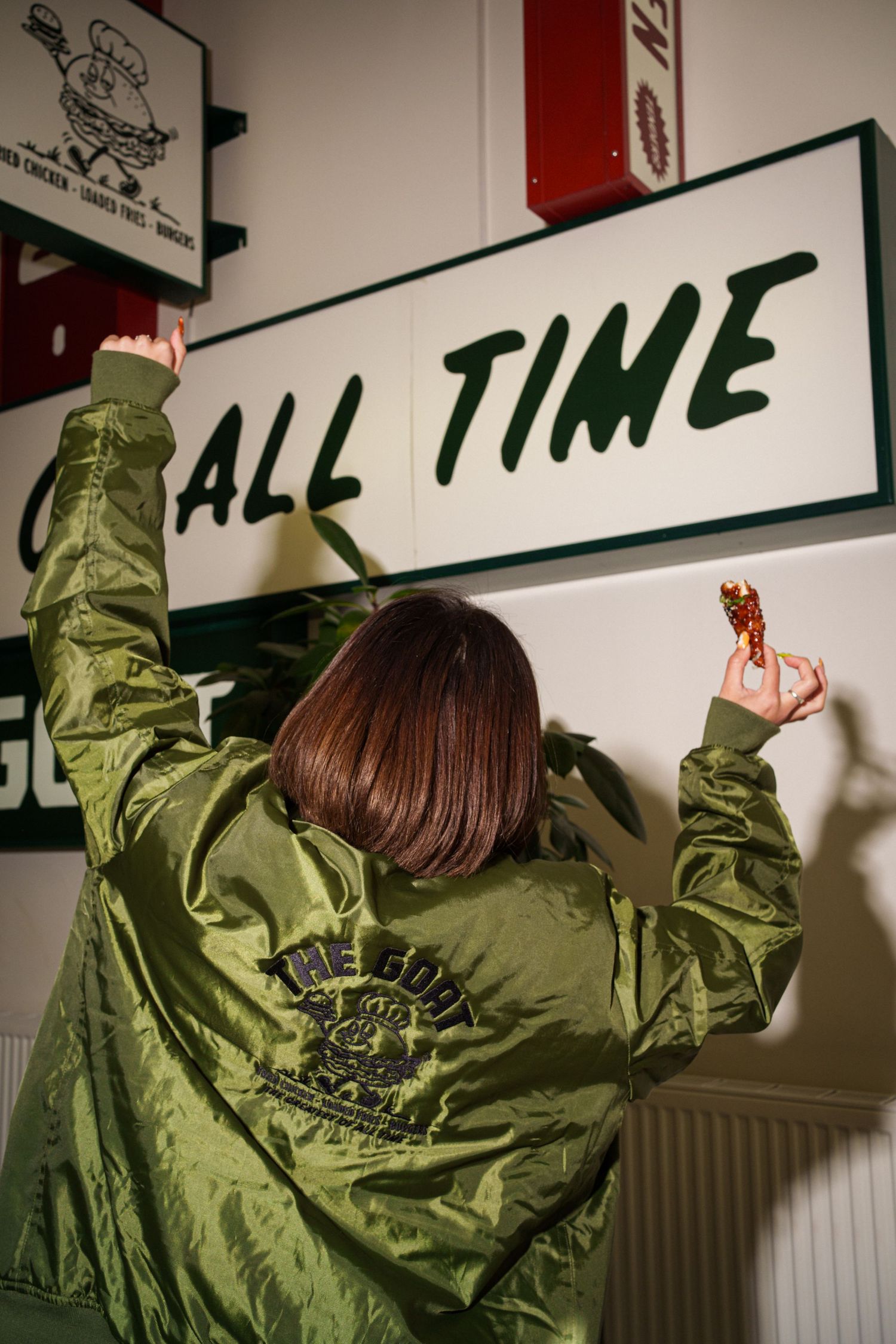 Person in olive green bomber jacket reaching up to touch an 'All Time' sign on a white wall.