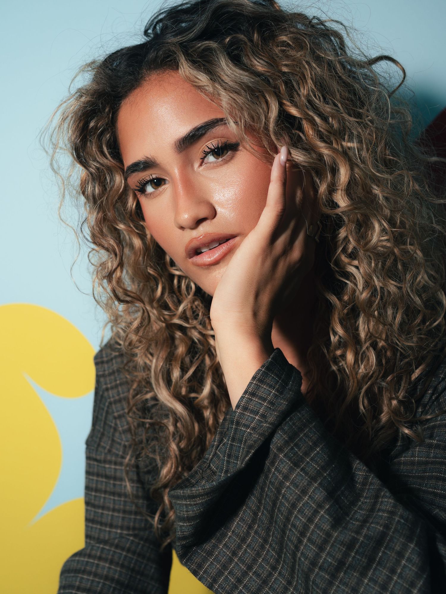 Woman with long curly highlighted hair posing with hand on cheek, wearing a plaid blazer against a blue background.