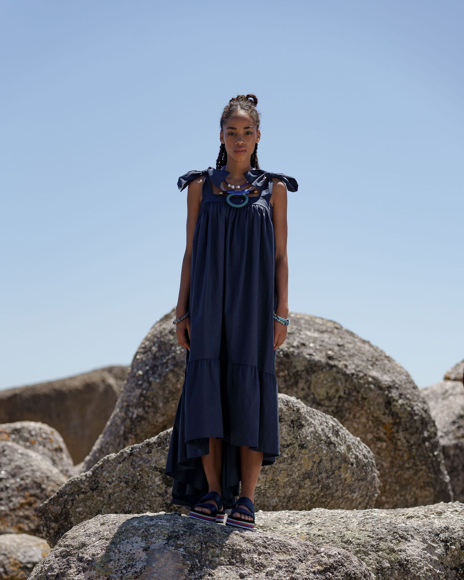 Woman in navy maxi dress with tie shoulders stands on rocks against blue sky.