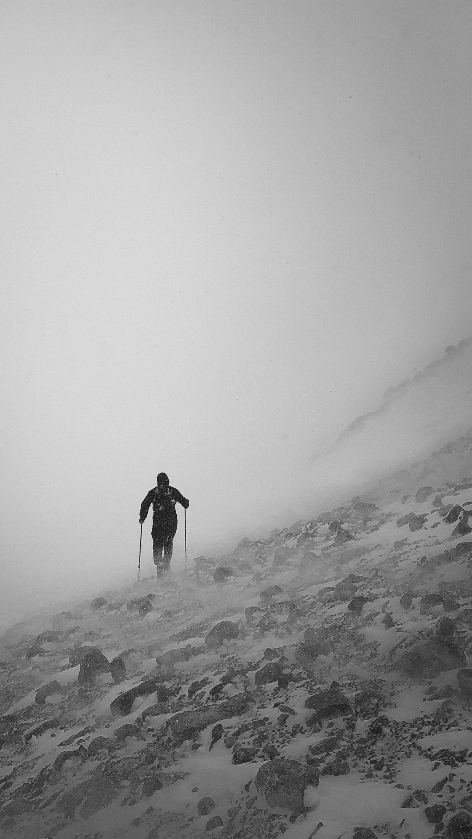 A lone hiker with trekking poles climbs a snowy, rocky mountain slope in dense fog, captured in black and white.
