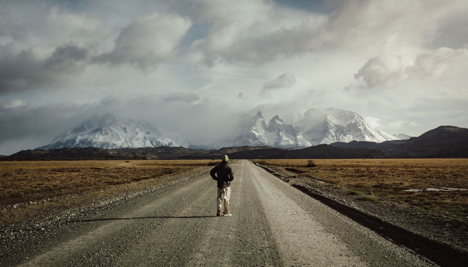 A lone hiker with a backpack walks down a straight road toward snow-capped mountains under dramatic cloudy skies.