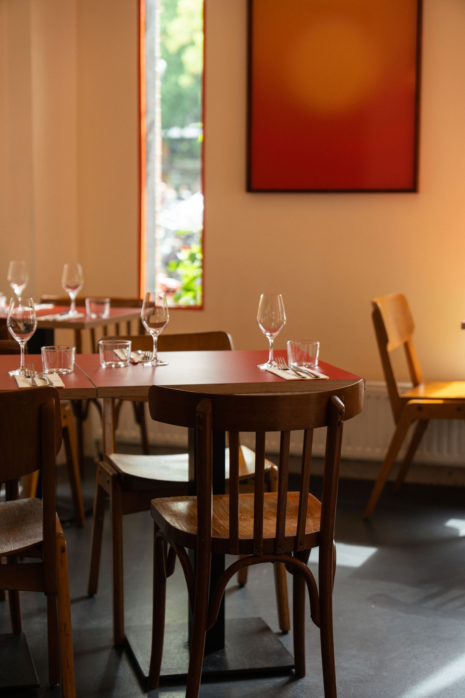 Cozy restaurant interior with round red tables, wooden chairs, wine glasses, and a large red abstract painting.