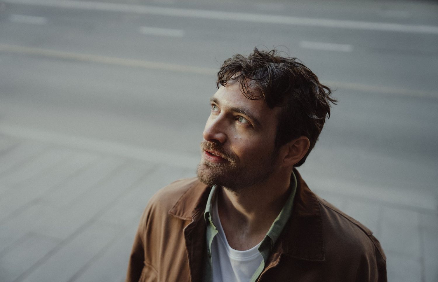 A person in a brown leather jacket and white t-shirt looks to the side in a moody indoor setting.