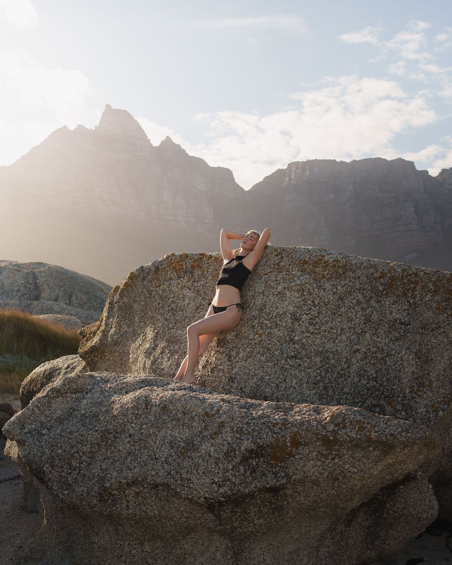 Woman in black bikini posing on large boulder with dramatic mountain peaks and golden sunlight in the background.