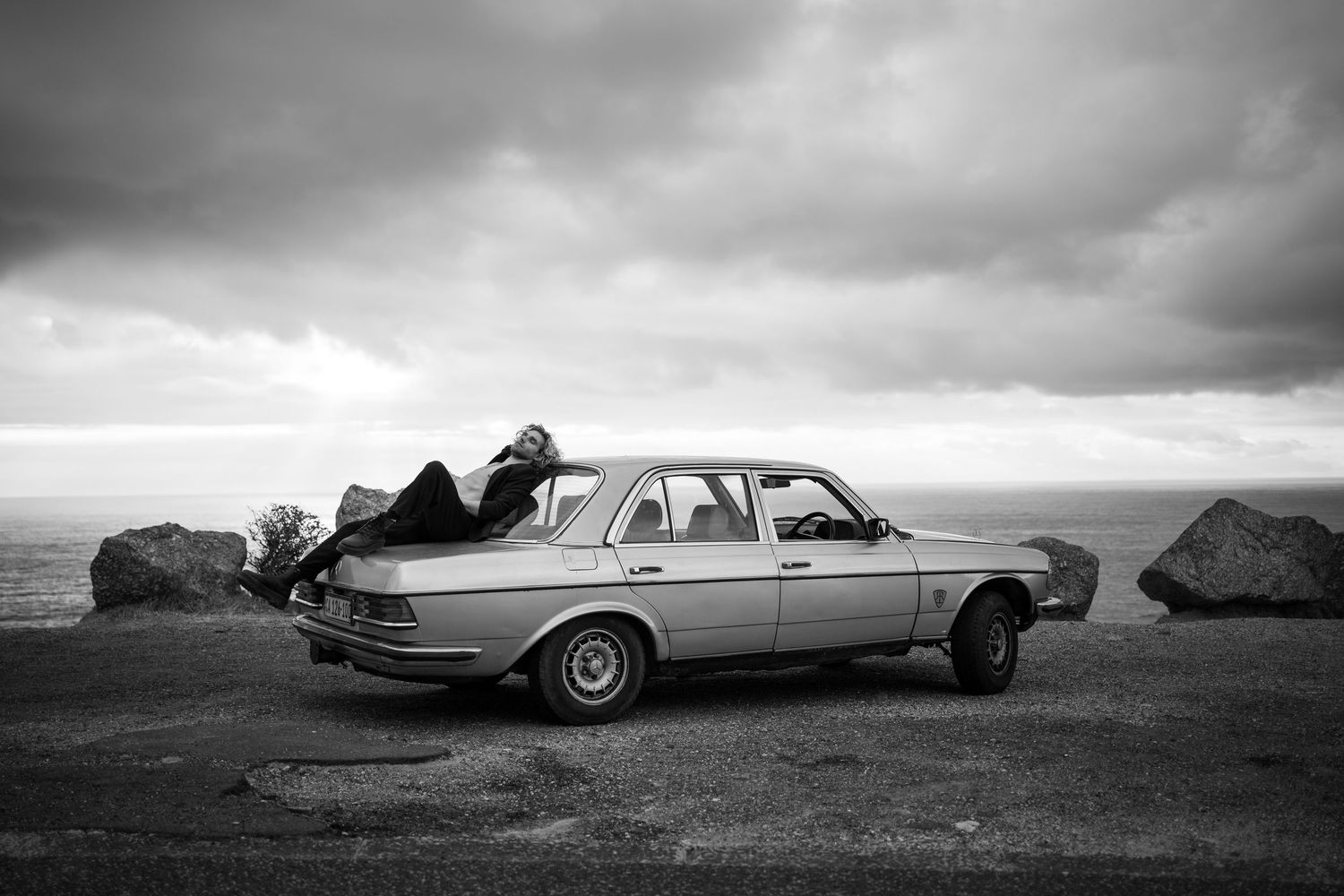 Person leaning into a vintage sedan parked on rocky coastal overlook under dramatic cloudy sky, black and white photo.