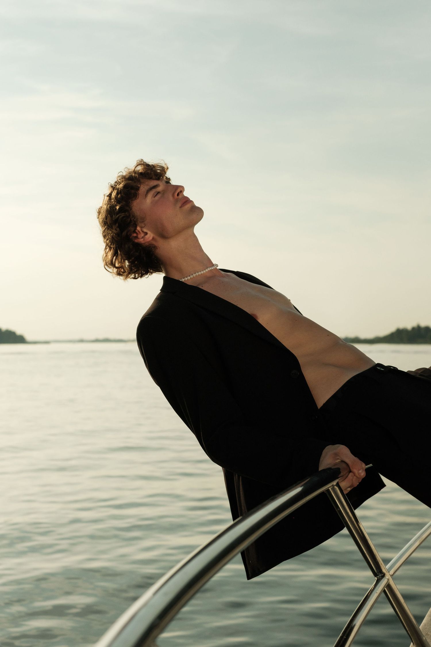 Young man with curly hair in open black blazer reclines on yacht bow, eyes closed, enjoying serene waterfront sunset.