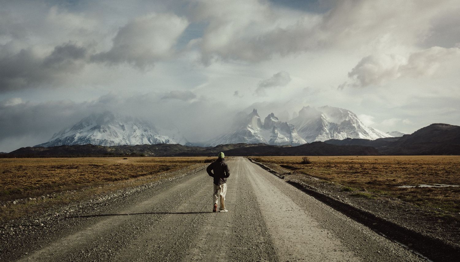 Long empty road leading to snow-capped mountains under dramatic cloudy skies.