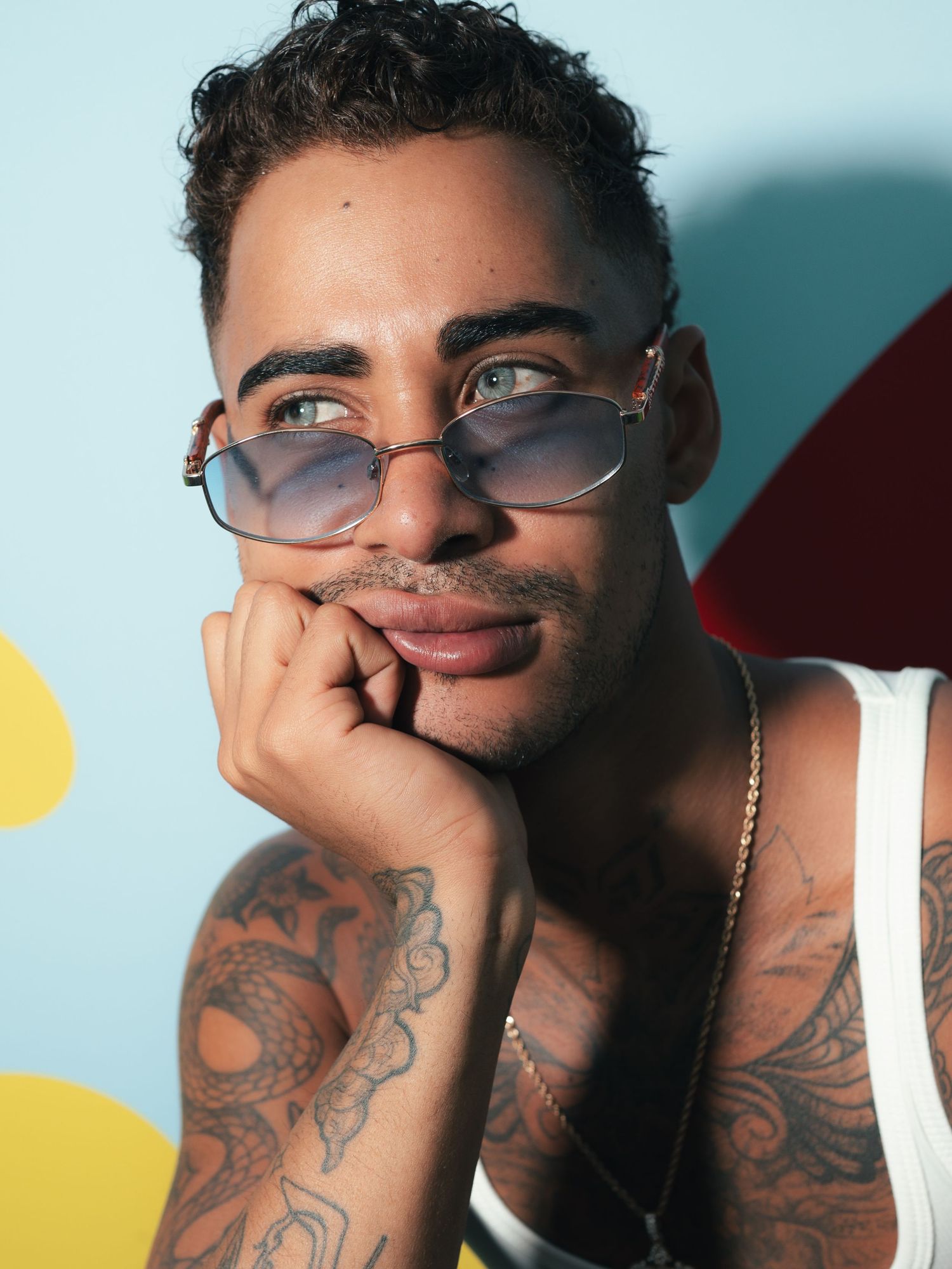 Young tattooed man with small tinted sunglasses resting chin on hand against colorful background.