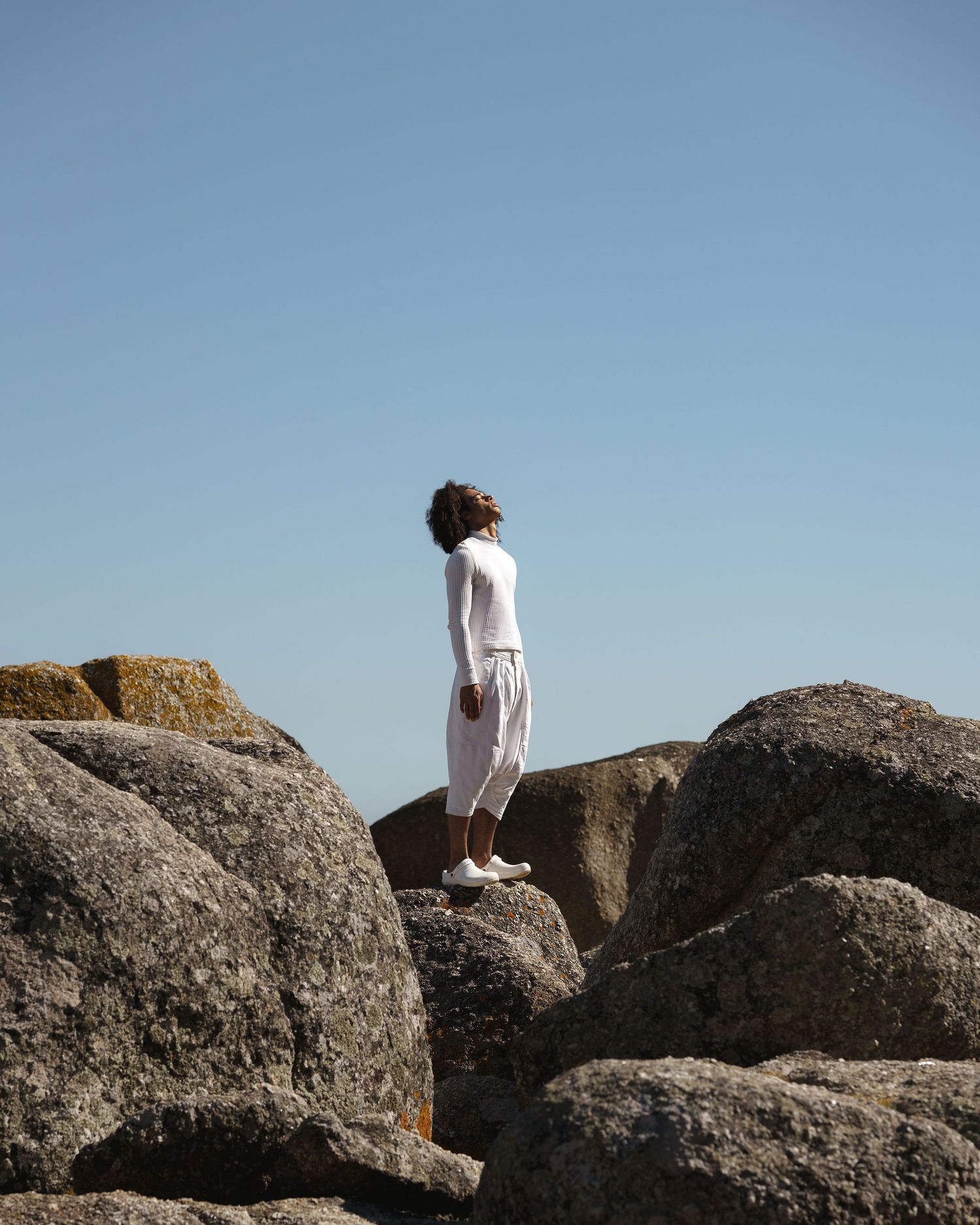 Person in white outfit standing on large coastal rocks against a clear blue sky.