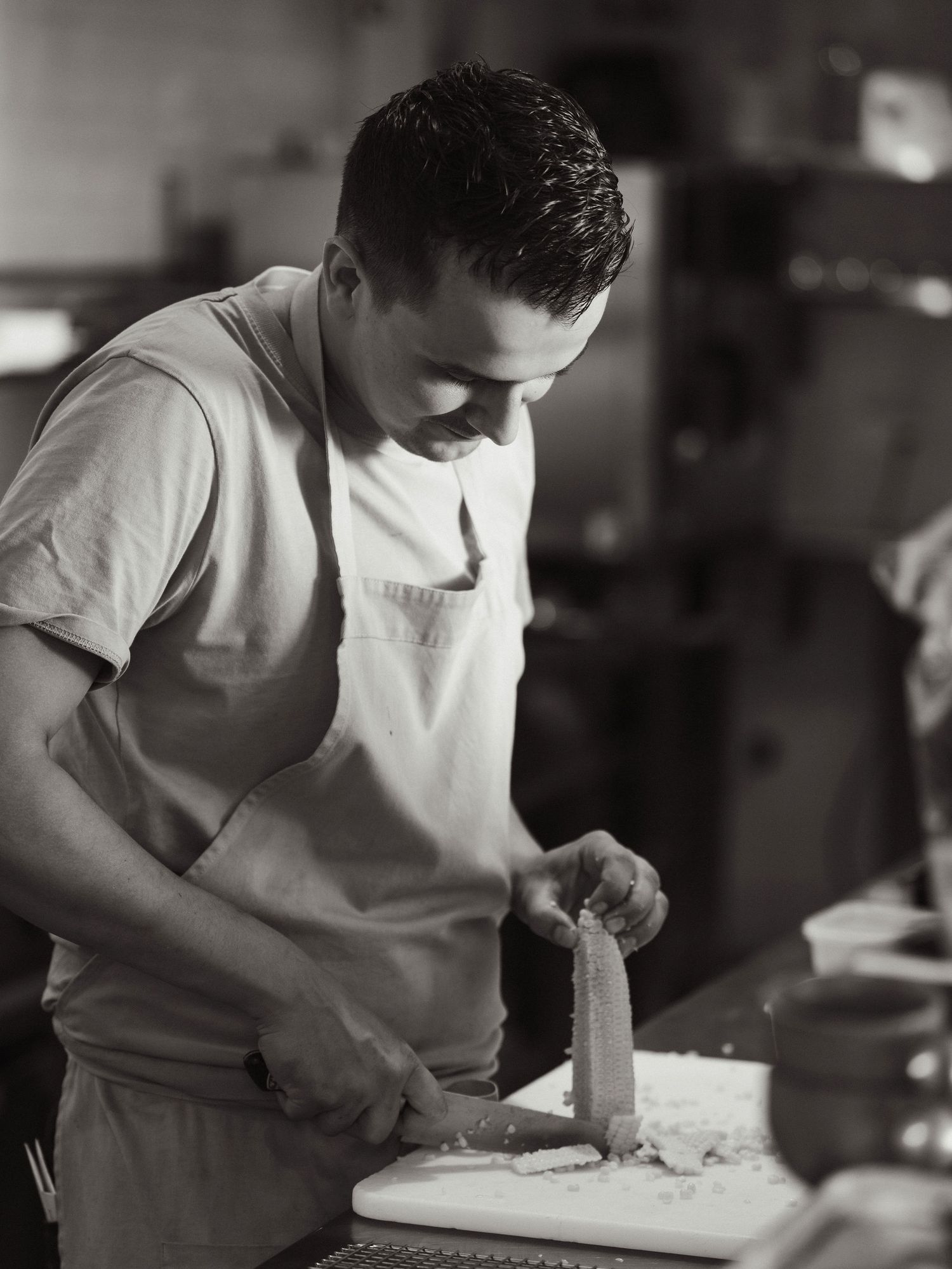 A chef in a white apron skillfully cuts food on a cutting board in a professional kitchen, captured in black and white.