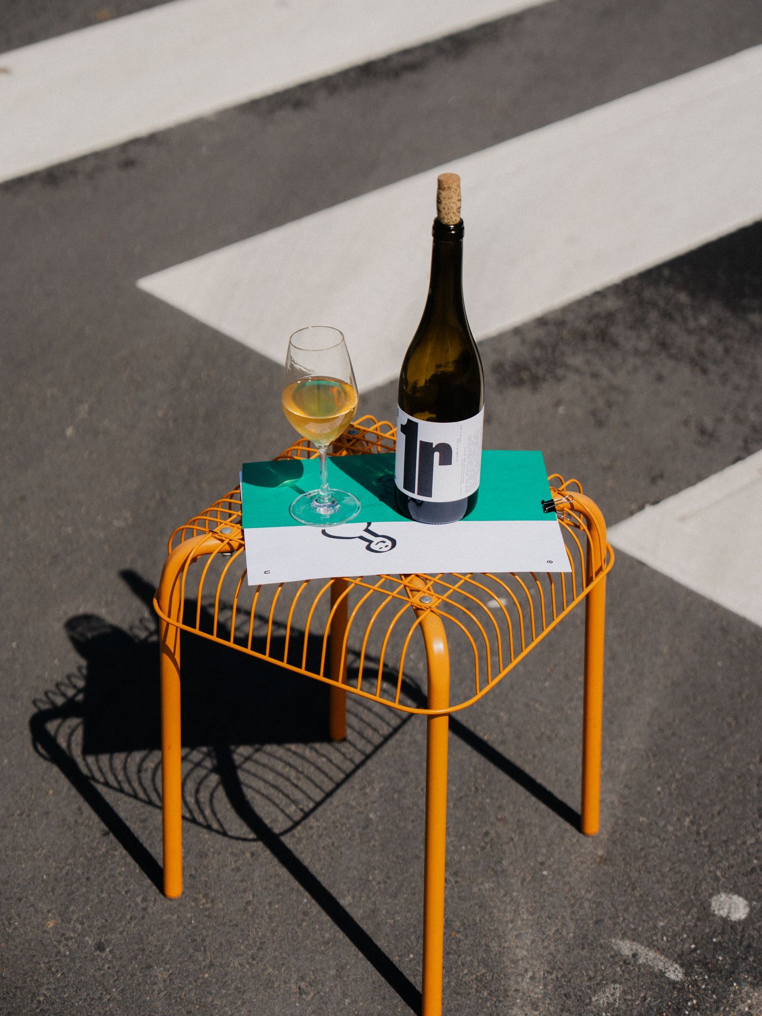 Bottle of white wine and glass on an orange wire side table outdoors on pavement.