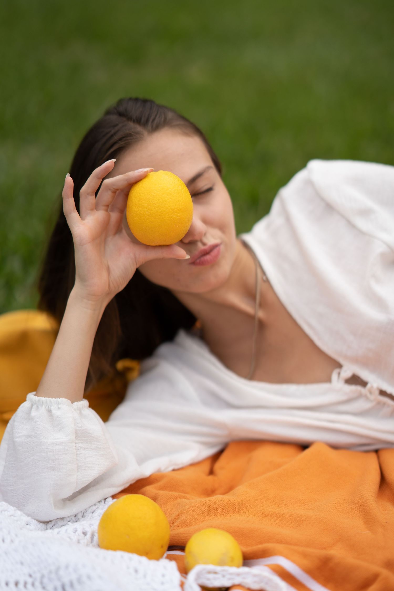 Young woman lying on grass holding a lemon over her eye, surrounded by fresh lemons on a white blanket outdoors.