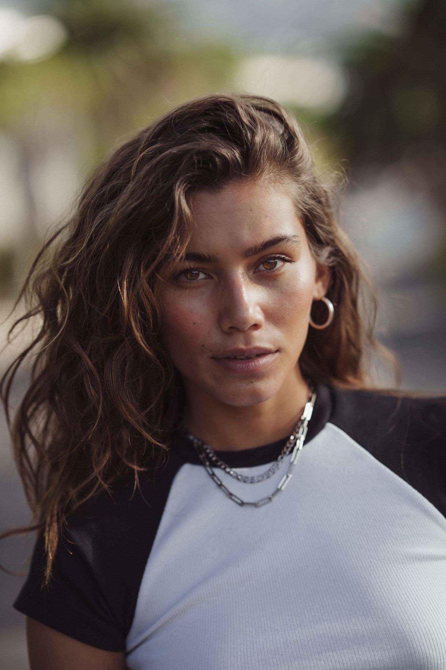 Young woman with wavy brown hair wearing a white and black top with silver chain necklaces, posing outdoors.
