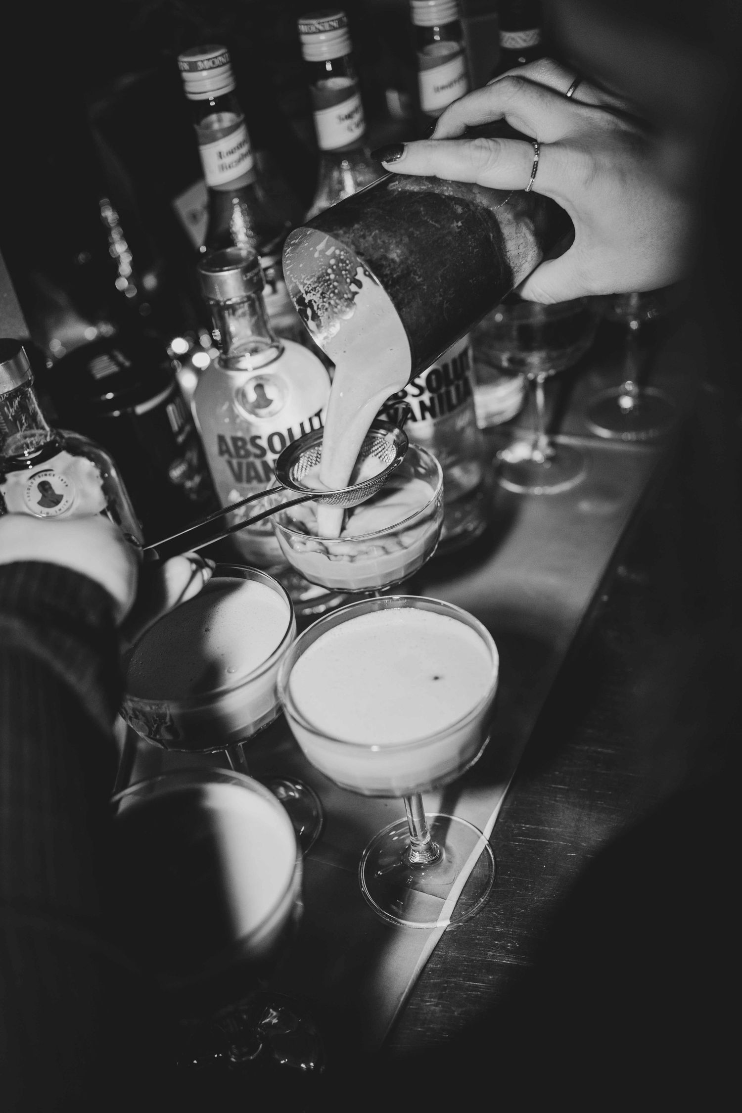 Bartender pouring cocktail into coupe glass at bar with Absolut Vodka bottles in black and white photo.