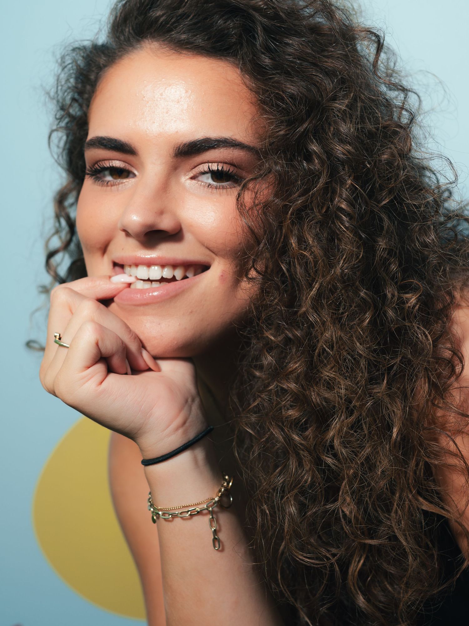 Smiling woman with curly brown hair, wearing a gold chain bracelet and ring, resting chin on hand against blue backdrop.