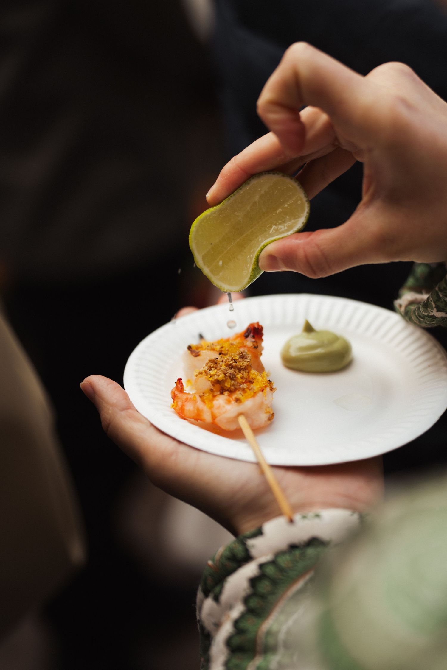 Hands squeezing fresh lime juice over a grilled shrimp skewer with green sauce on a white paper plate.