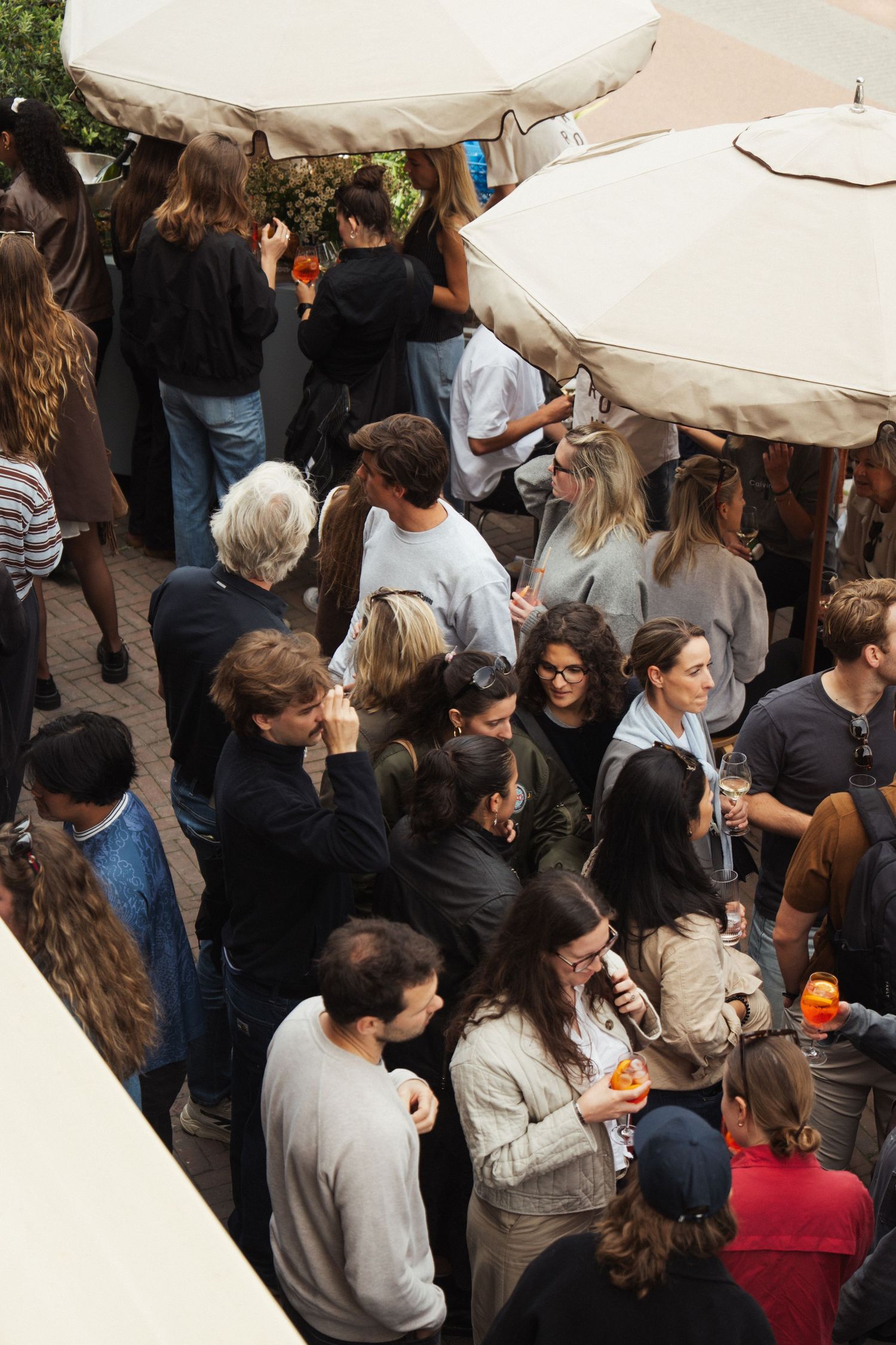 Crowd of people socializing outdoors under white umbrellas at a busy social gathering or street event.