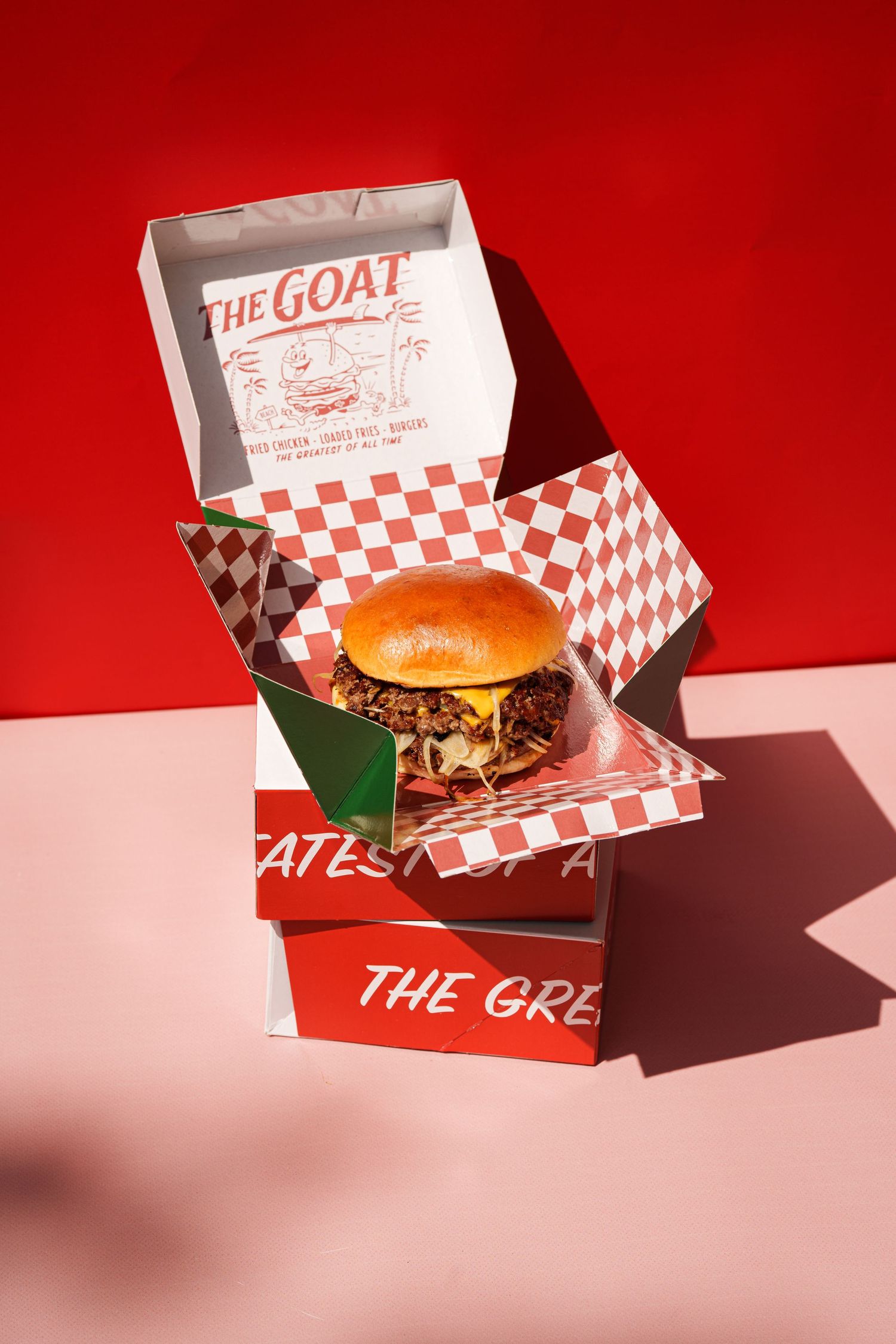 A juicy burger in a red checkered box from The Goat restaurant, set against a vibrant red background.