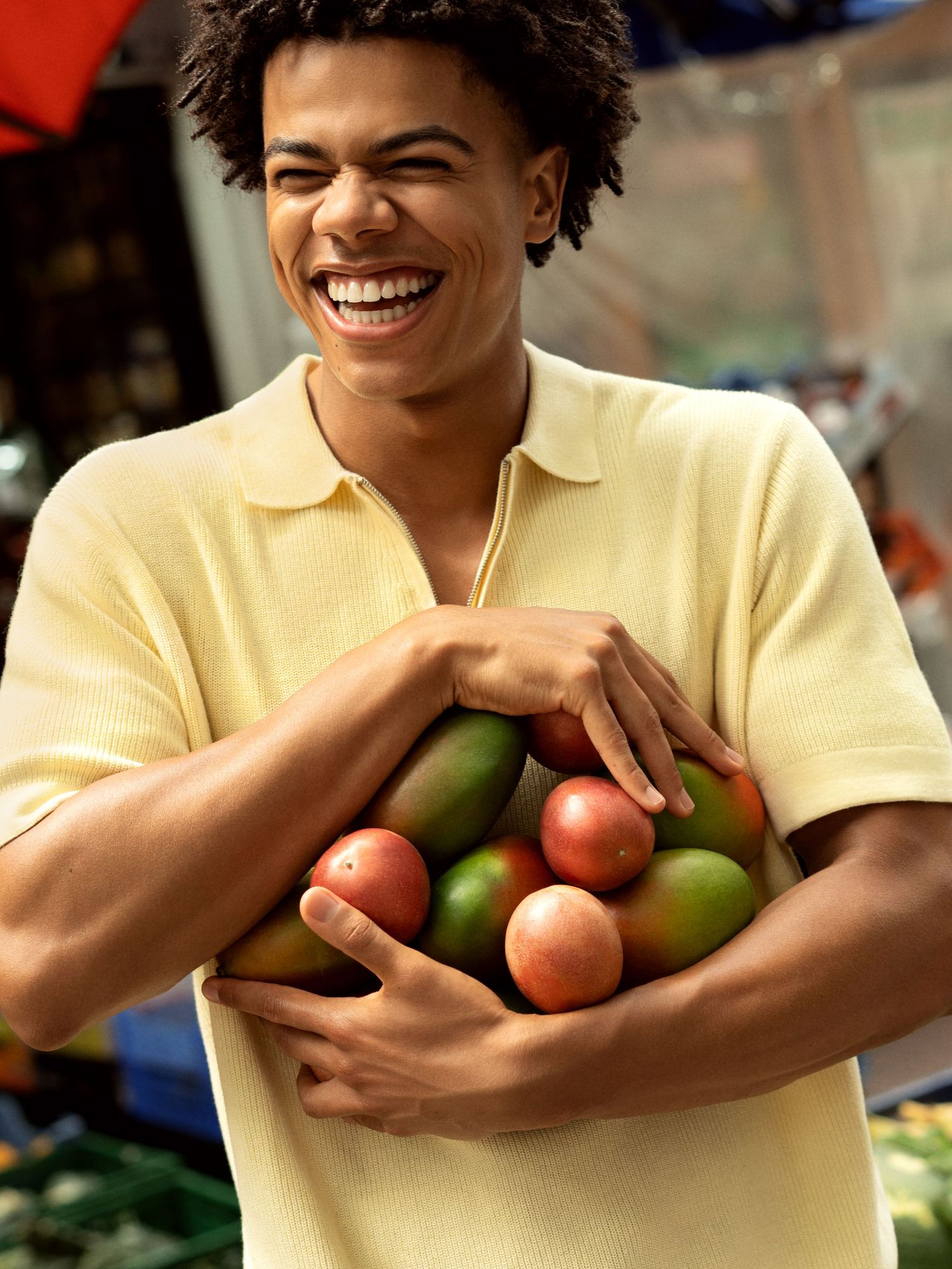 Smiling young man holding an armful of fresh mangoes and tomatoes at an outdoor market.
