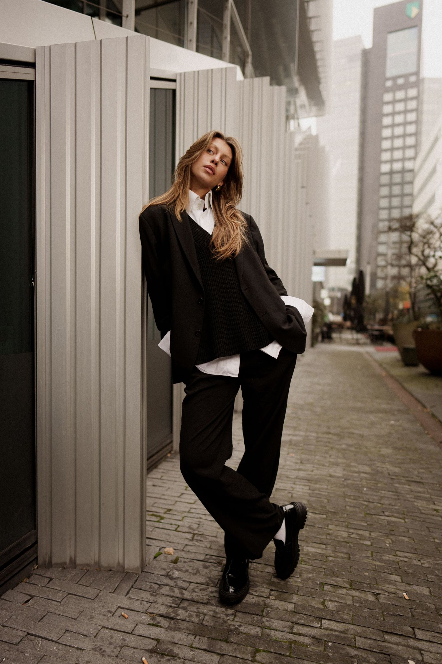 Woman in black oversized blazer, trousers, and loafers leans against a building on a city street.