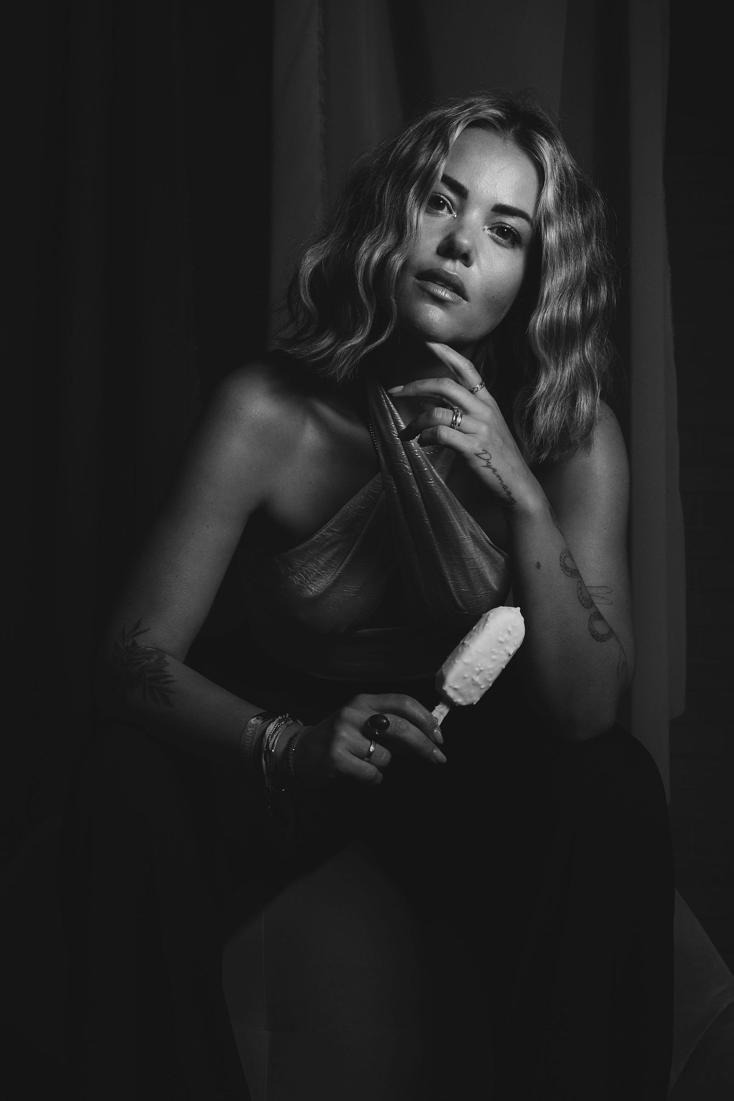 Black and white portrait of a woman with wavy hair holding a brush, tattoos visible, posing against dark curtains.