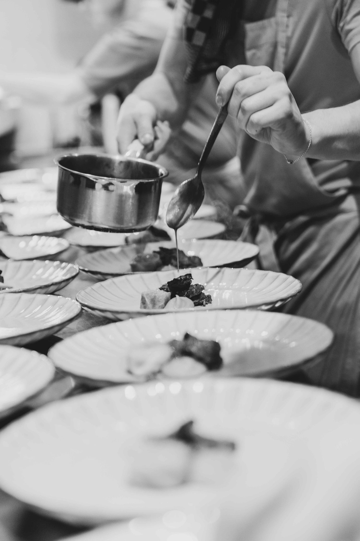 Black and white photo of a chef plating gourmet dishes, pouring sauce from a pan onto multiple white plates.
