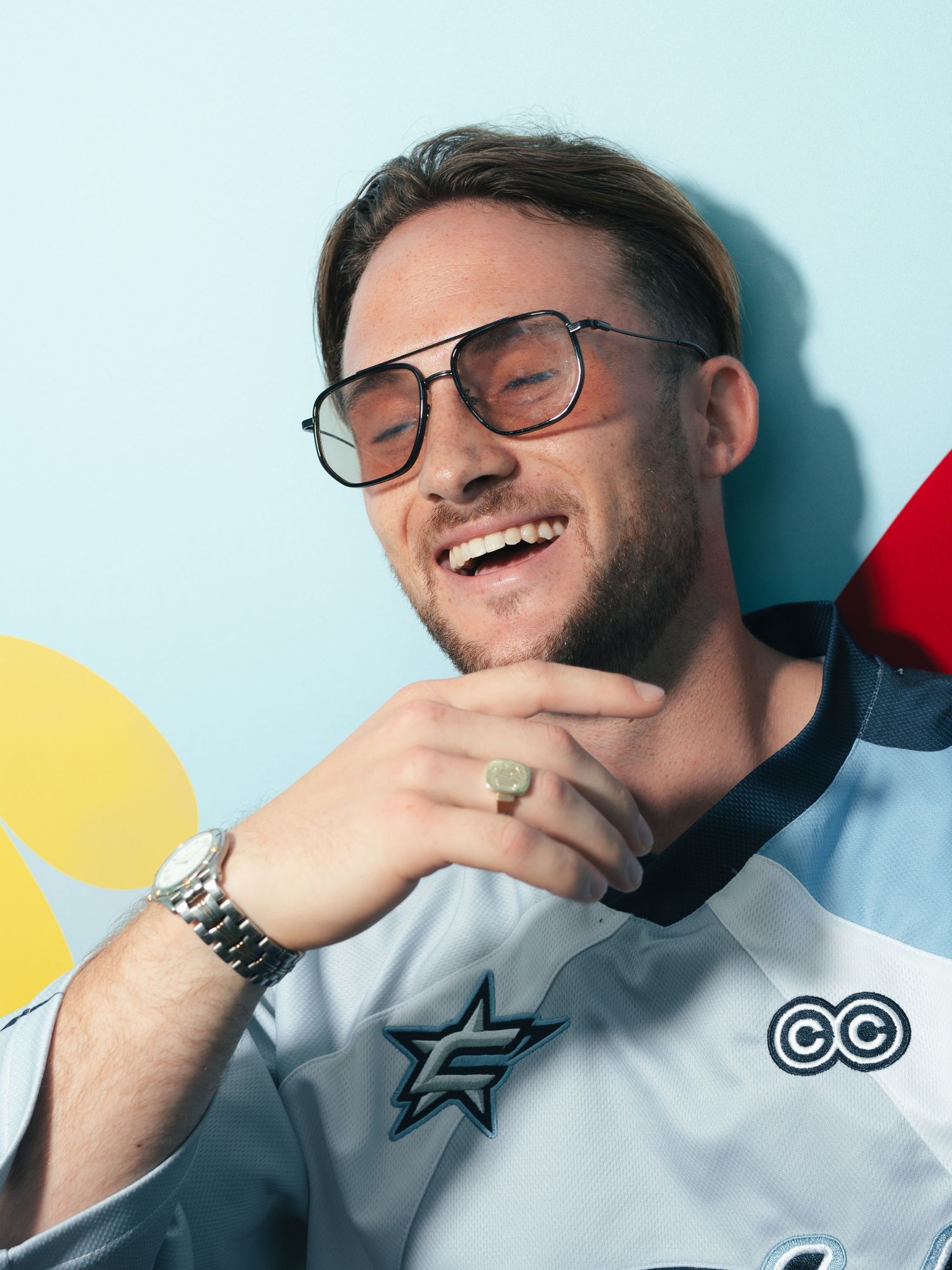 Laughing man in grey jersey wearing aviator sunglasses and silver ring watch, posing against light blue background.