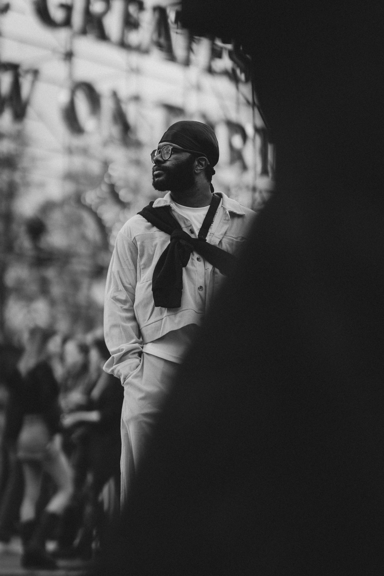 Black and white photo of a stylish man in glasses, beanie, and white outfit with scarf, standing outdoors at a busy event.