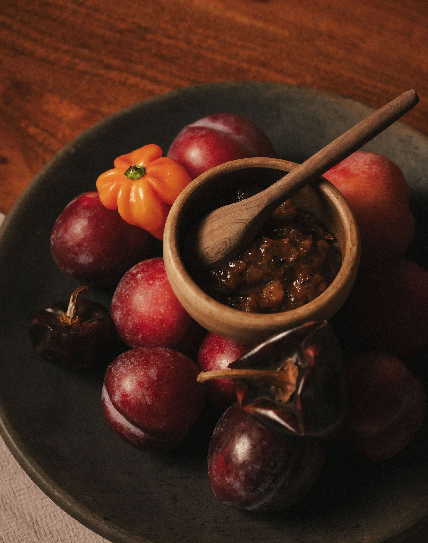 Dark plate with plums, orange tomato, and a wooden bowl of fruit chutney with spoon on rustic table.