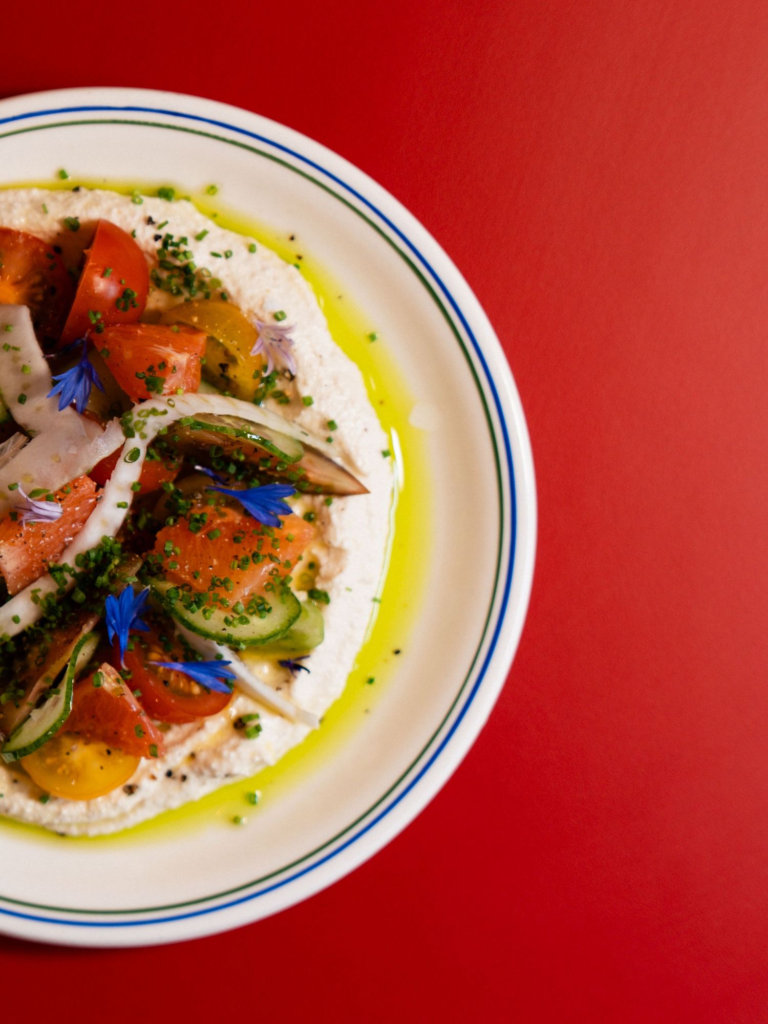 Fresh heirloom tomato salad with whipped ricotta, cucumber, olive oil and edible flowers on white plate.
