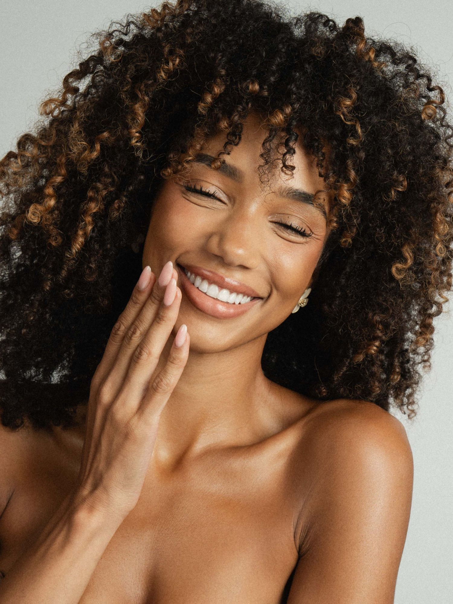 Smiling young Black woman with voluminous curly hair gently touching her glowing skin against a gray background.