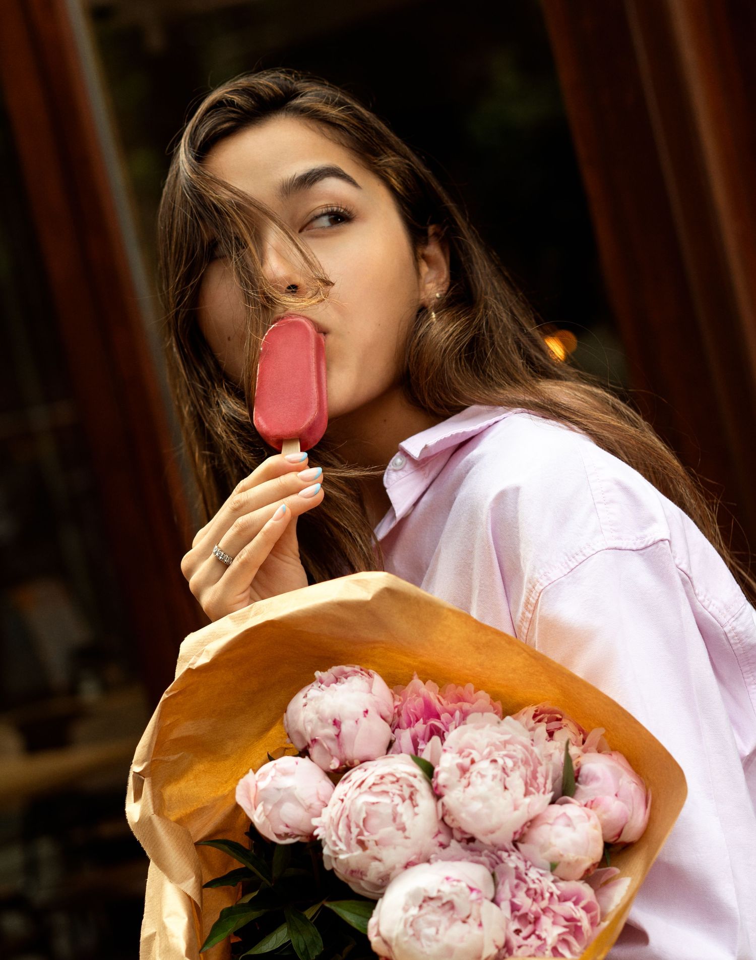 Young woman licking a pink ice cream bar while holding a bouquet of pink peonies wrapped in brown paper.