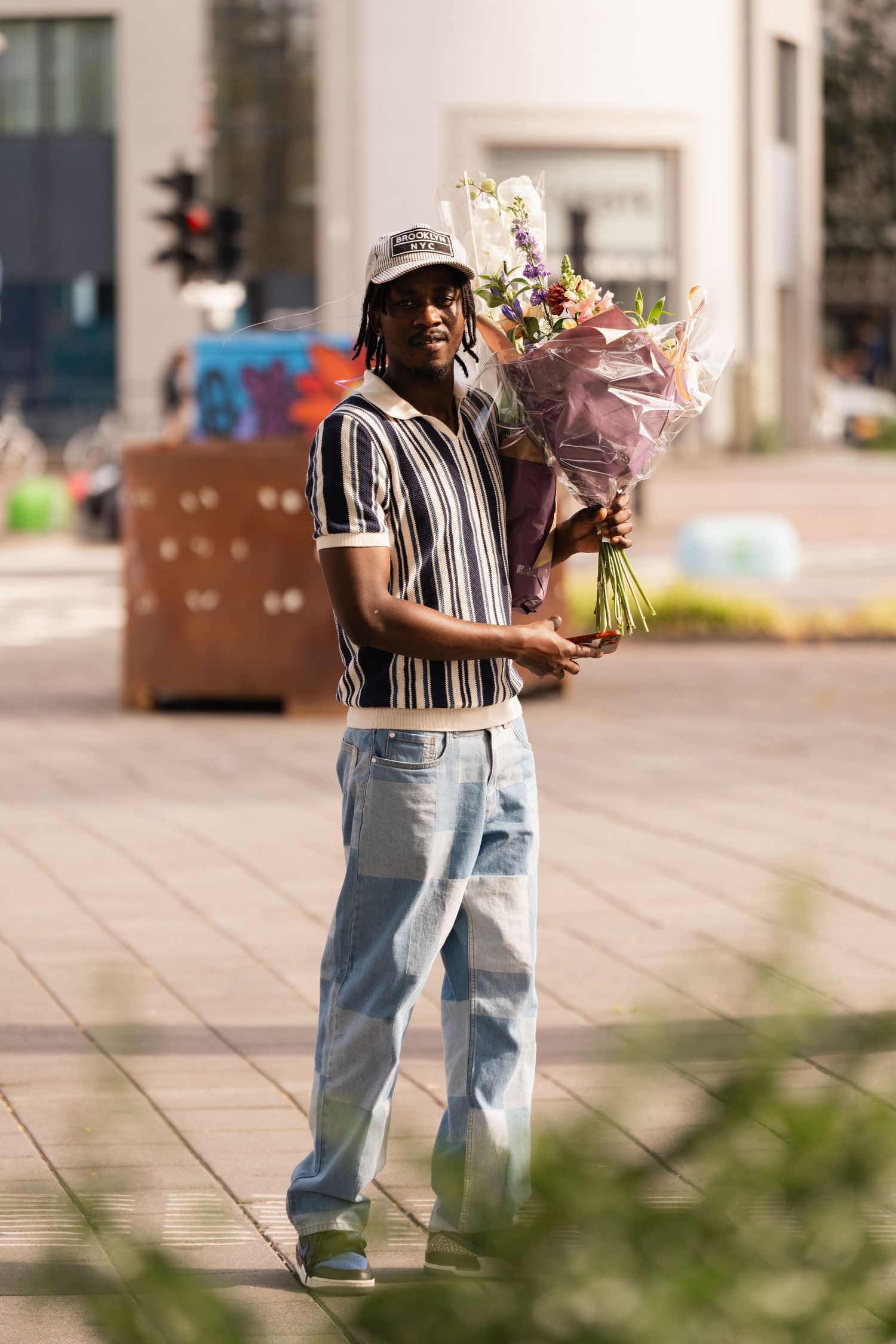Young man in striped shirt and jeans holding a bouquet of flowers on a sunny urban street.