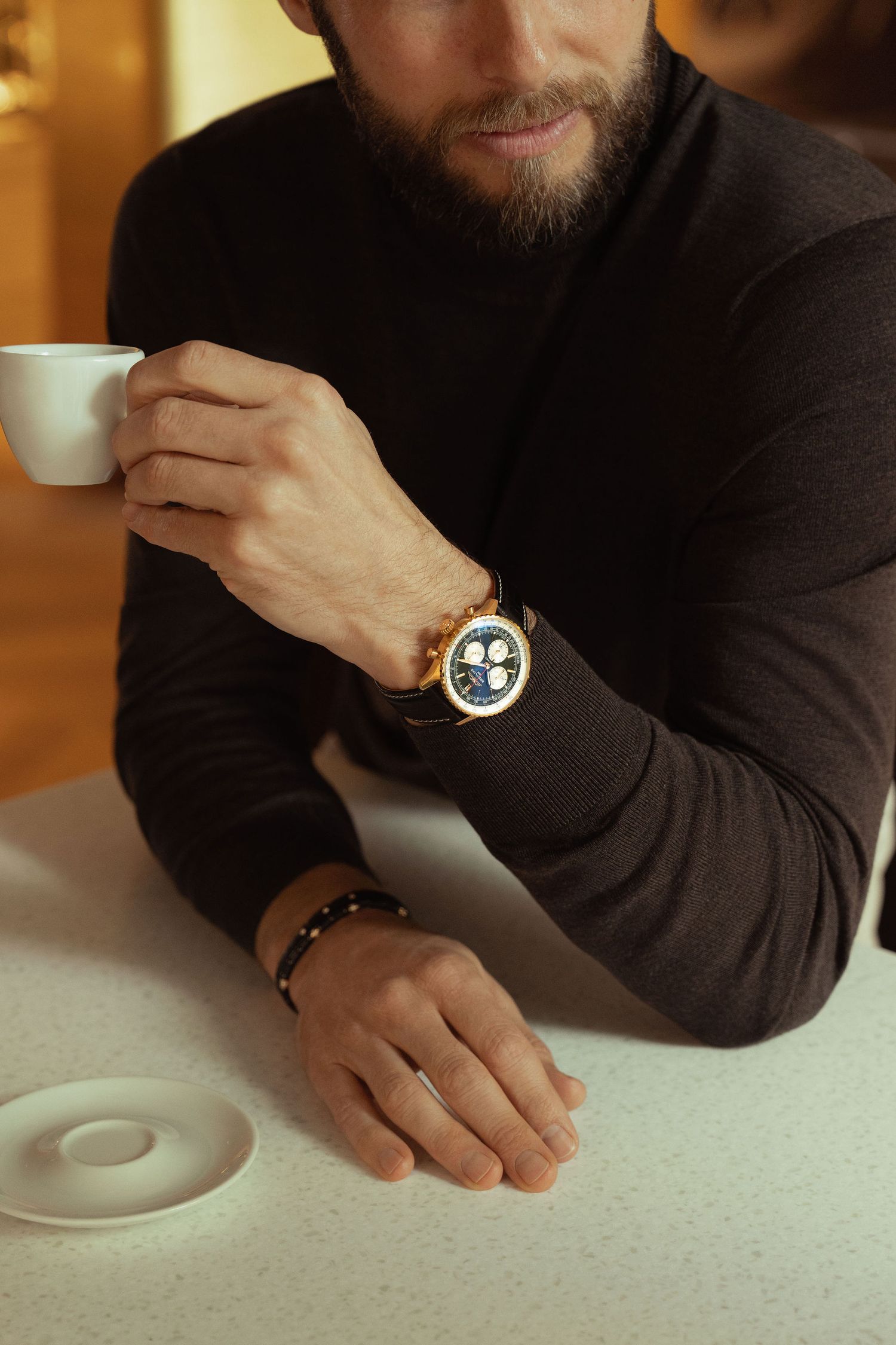 Bearded man in black turtleneck wearing luxury chronograph watch, holding espresso cup at white table.