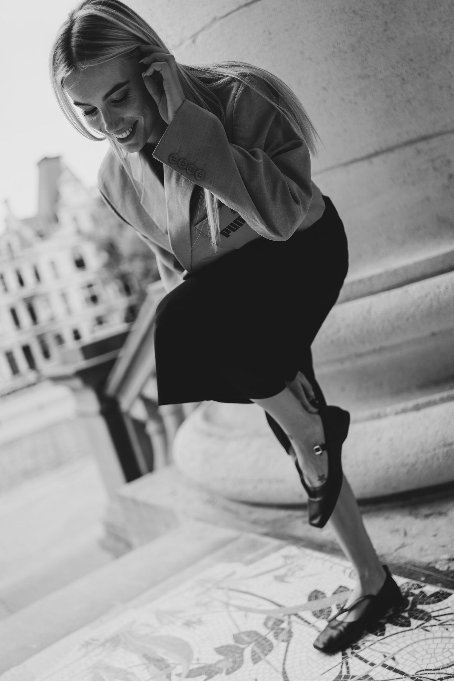 Smiling blonde woman in black midi skirt and white blouse laughing outdoors on steps, black and white photo.