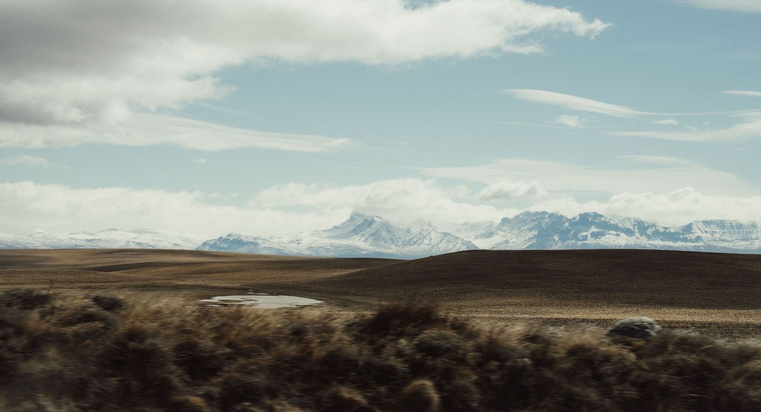 Vast Icelandic tundra landscape with snow-capped mountains, dramatic cloudy sky, and a small pond in the foreground.