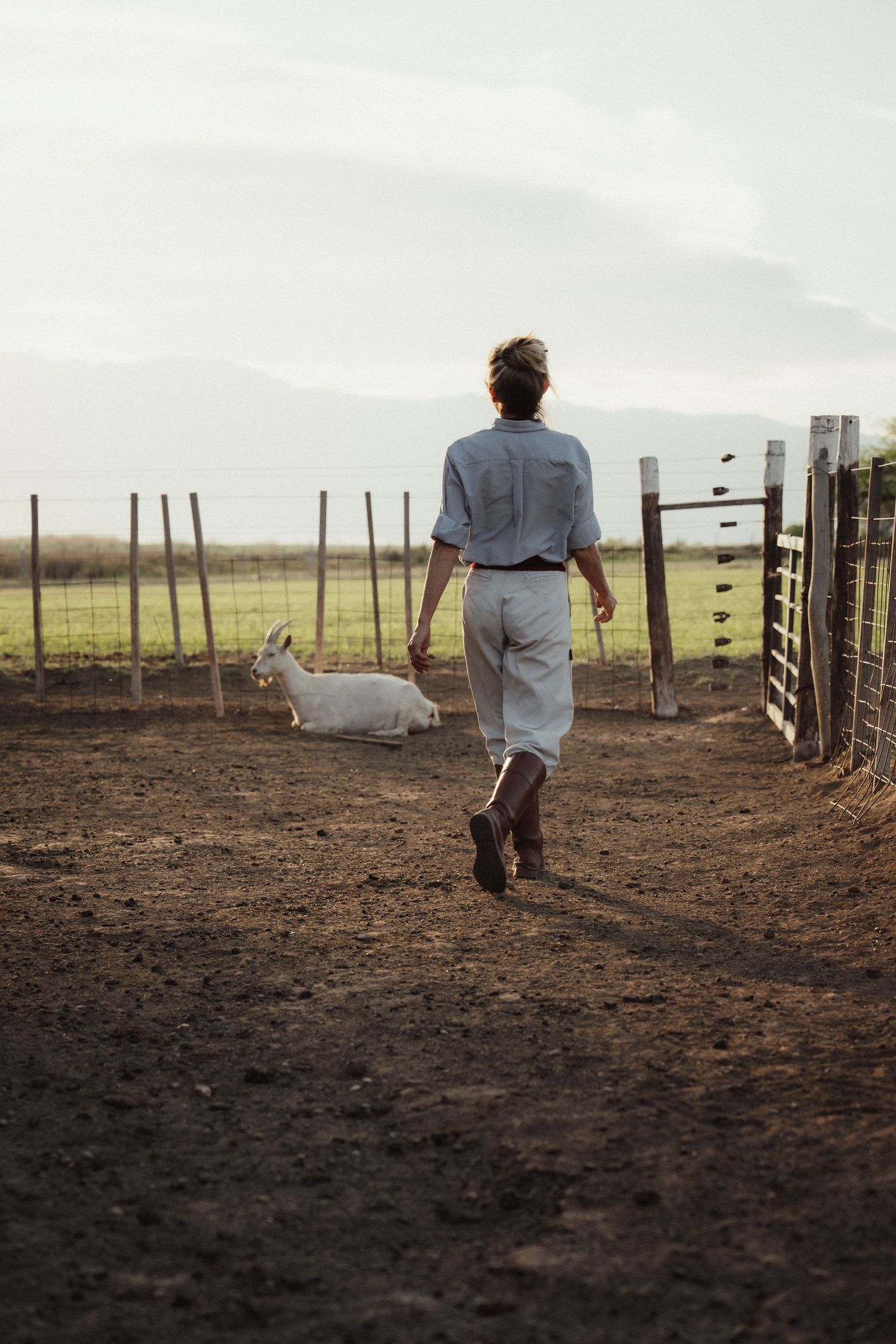 Figure in light clothing walks across a dusty corral with wooden fencing.