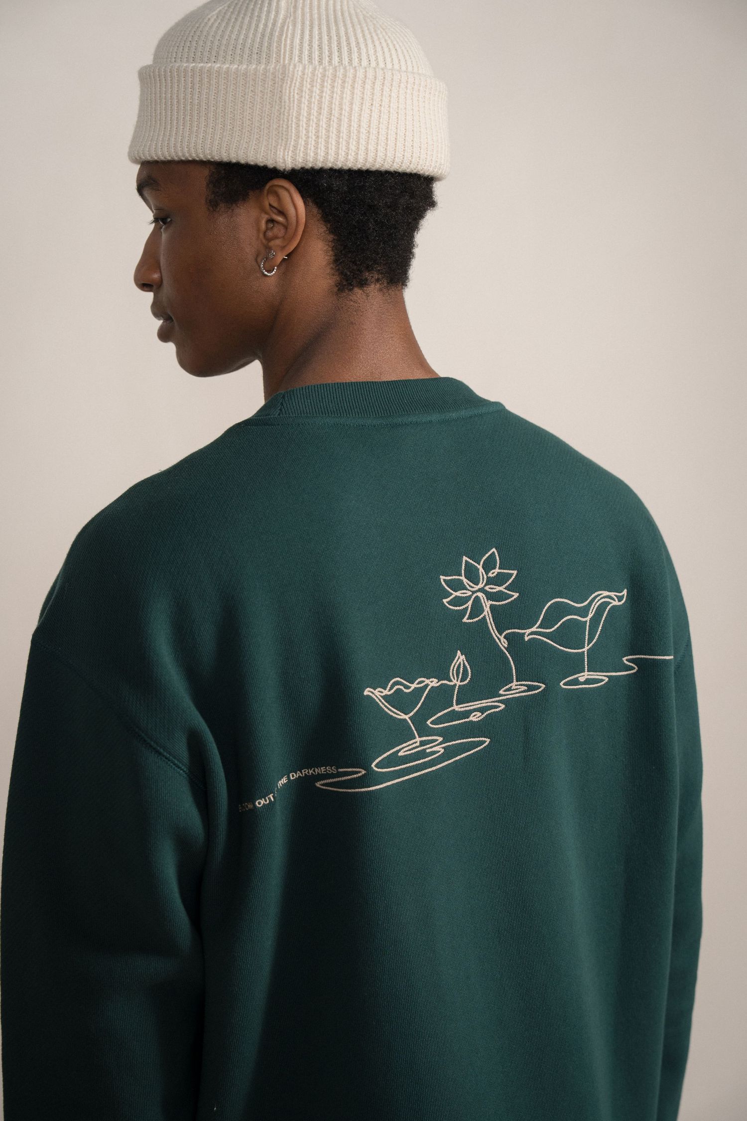Person wearing a white beanie and forest green sweatshirt with minimalist lotus flower and bird design on the back.